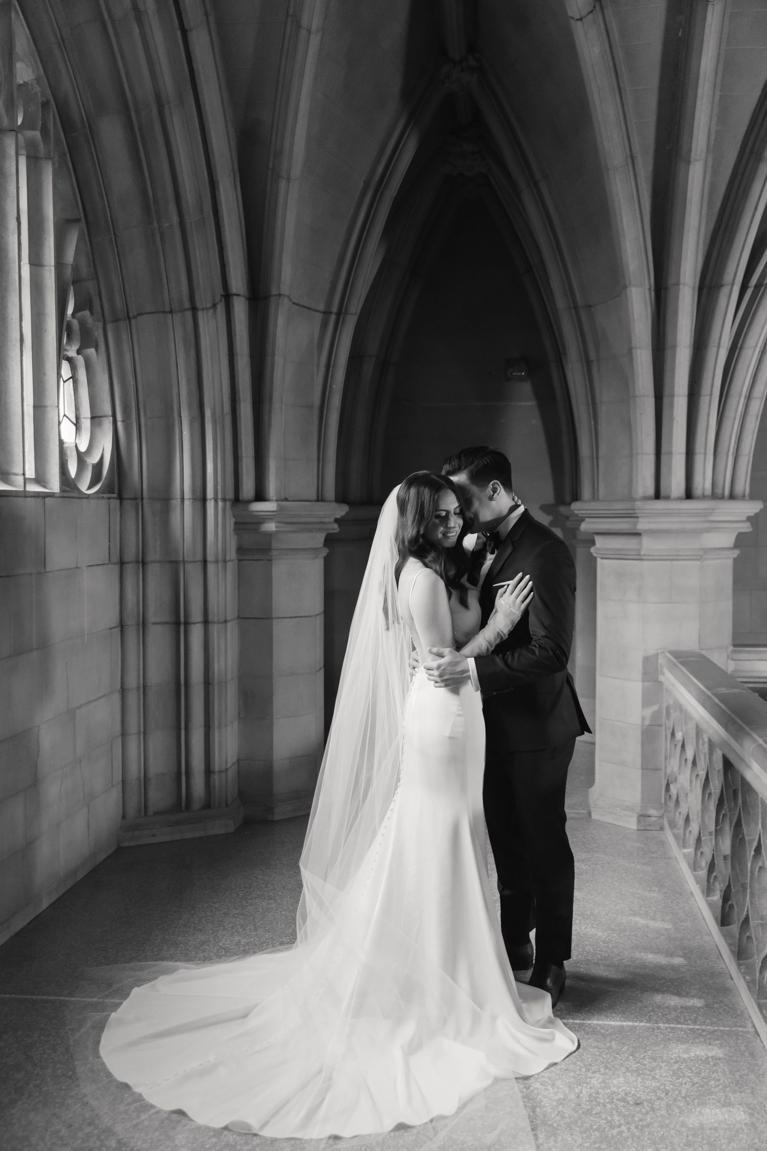Black and white wedding photo showing a romantic moment in a cathedral hallway with dramatic arches.