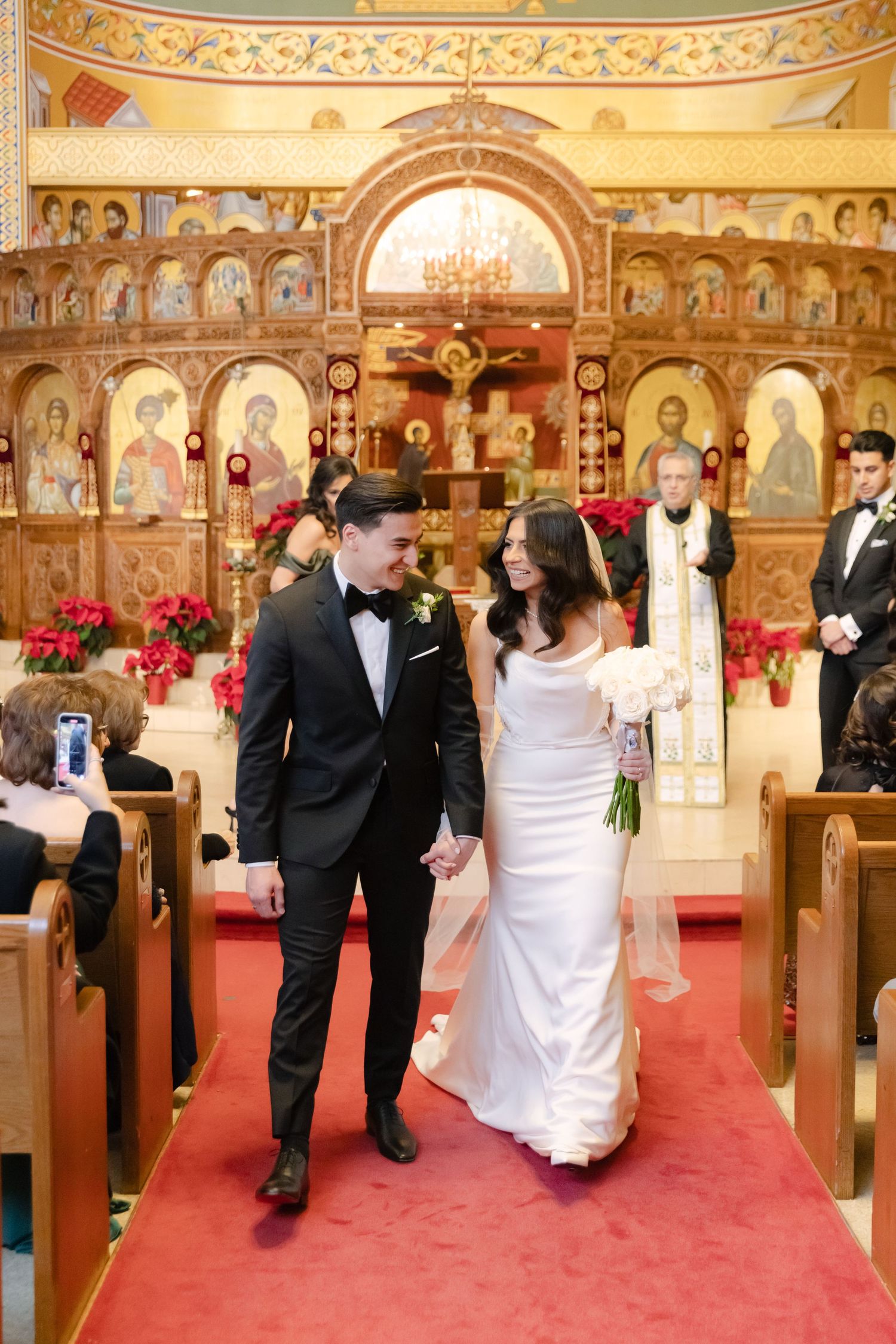 Newlyweds walk down red-carpeted aisle in Orthodox church with decorated iconostasis behind them.