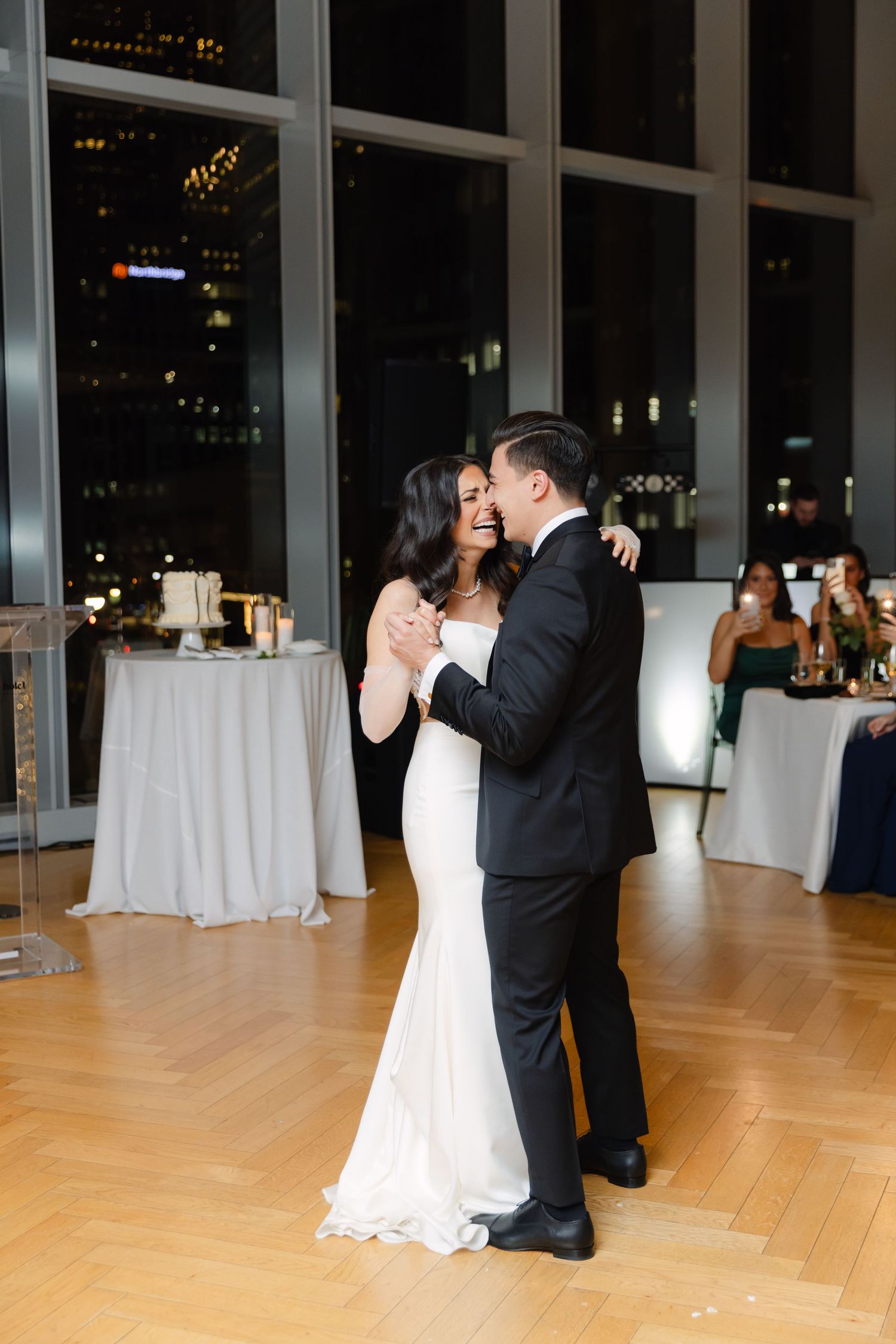 A couple shares an intimate dance moment at their wedding reception with city lights in the background.