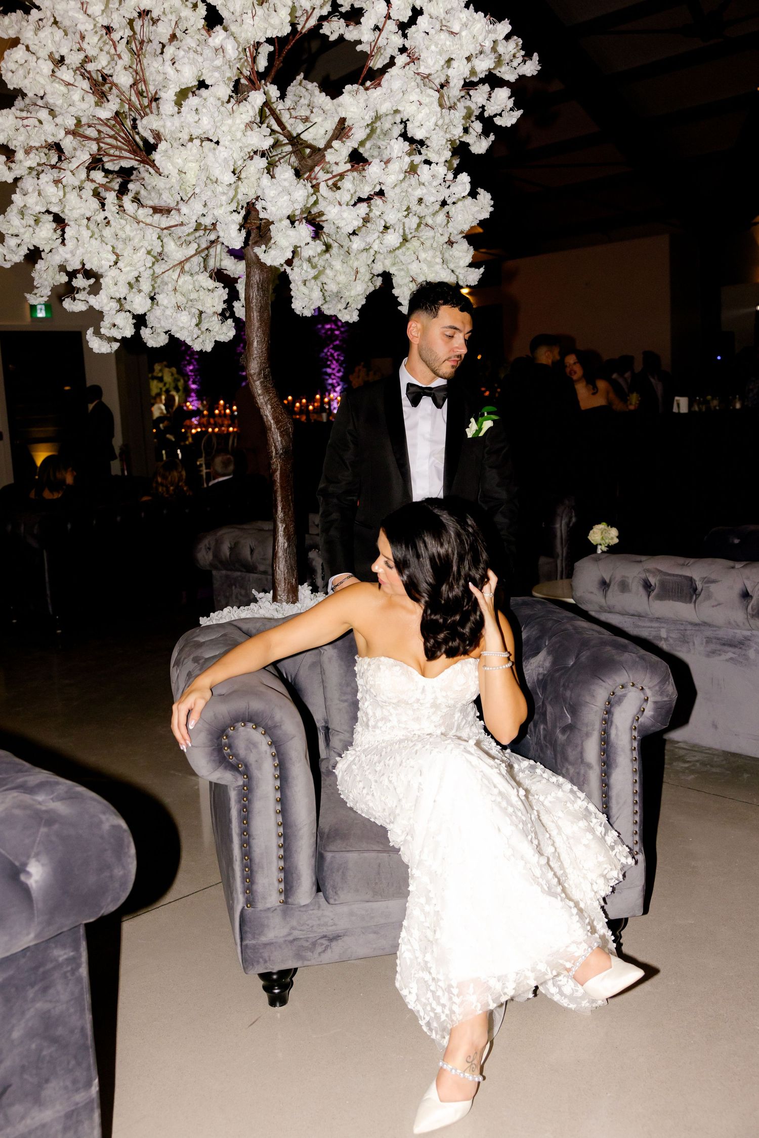Couple interacting playfully on elegant gray sofa beneath white floral wedding decor.