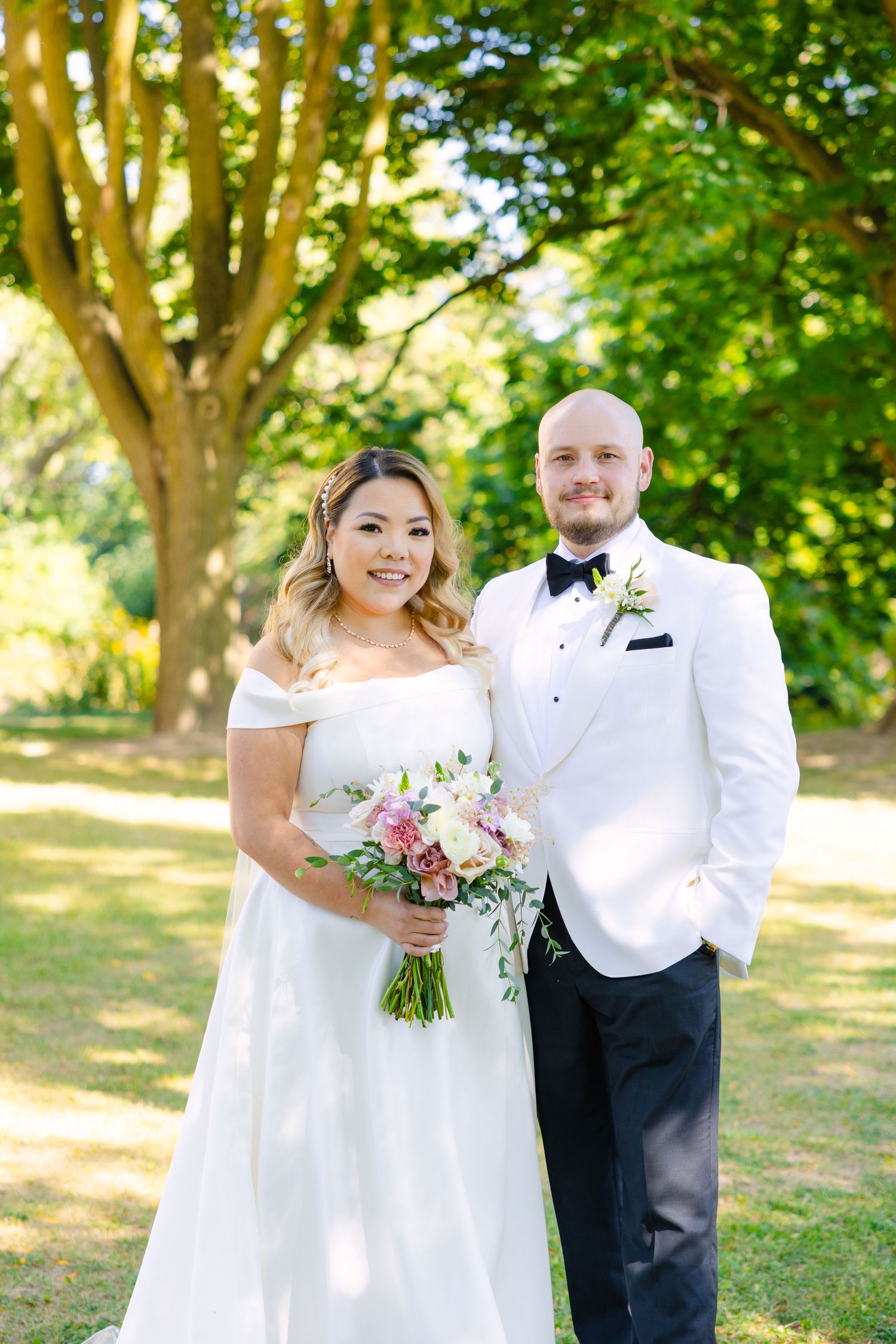 Elegant wedding portrait of couple in white attire standing in front of lush green trees.