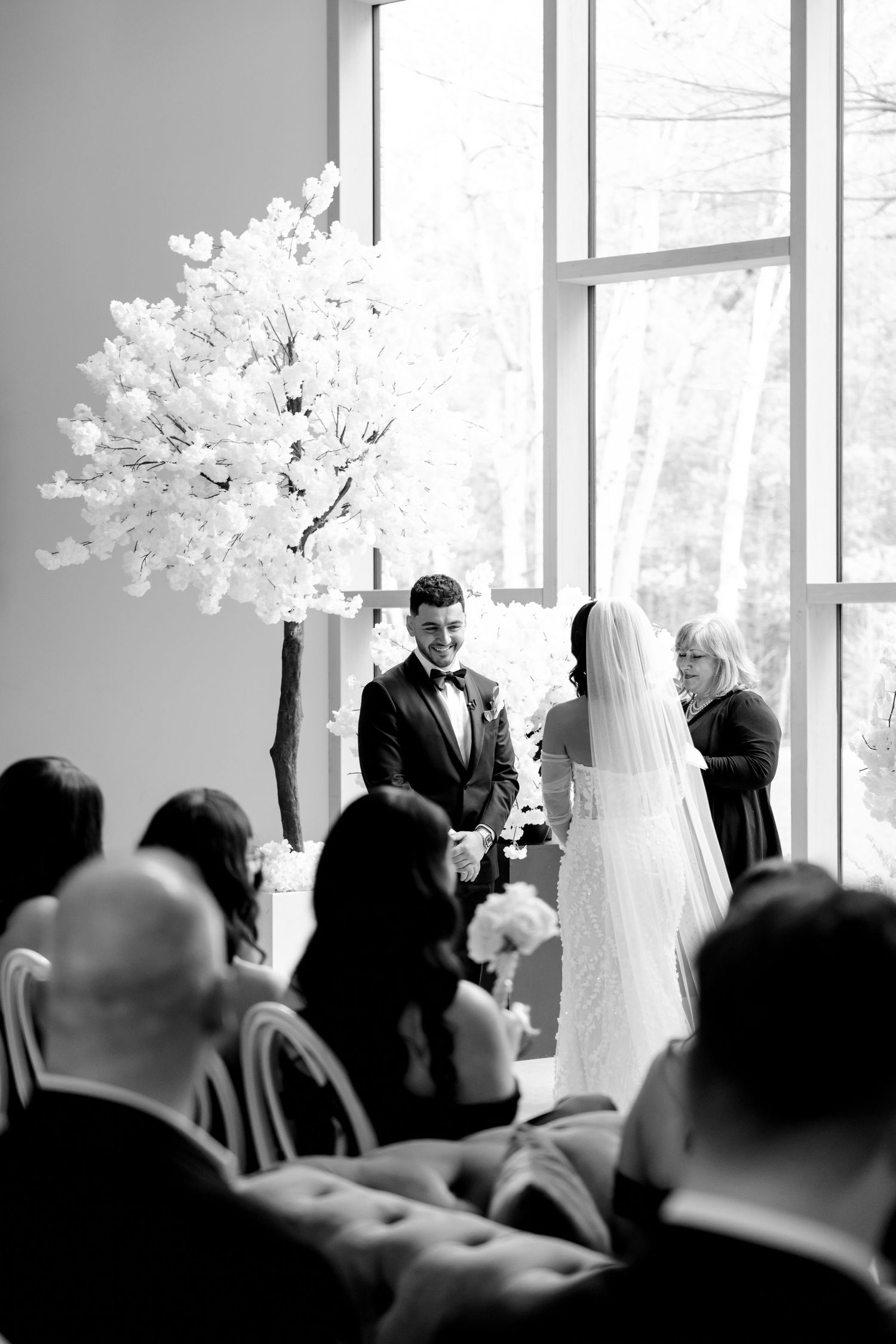 A black and white wedding ceremony photo featuring a large white floral arrangement and seated guests.