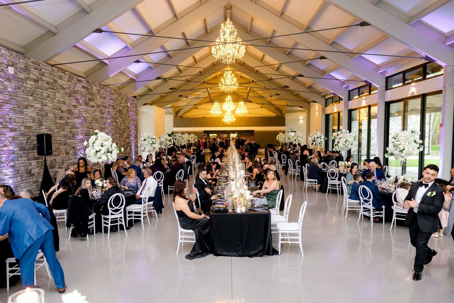 Spacious wedding reception hall with guests seated at round tables under crystal chandeliers and stone walls.
