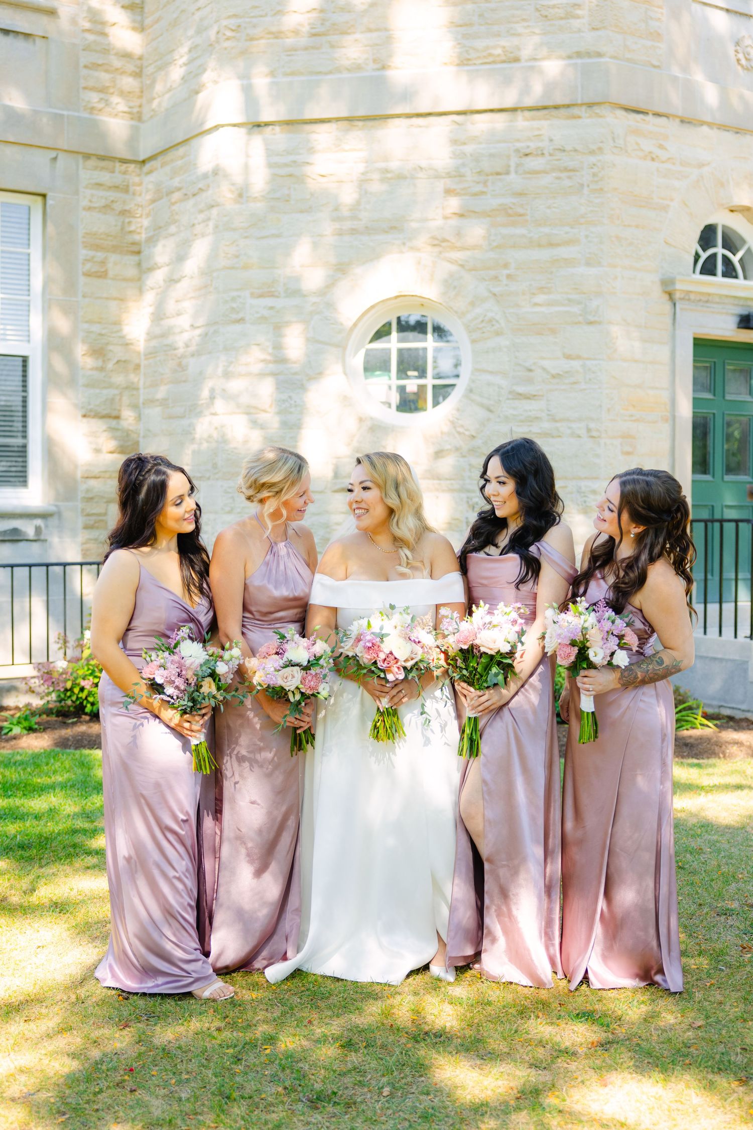 Bridal party in dusty rose dresses gather around bride for outdoor wedding photo.