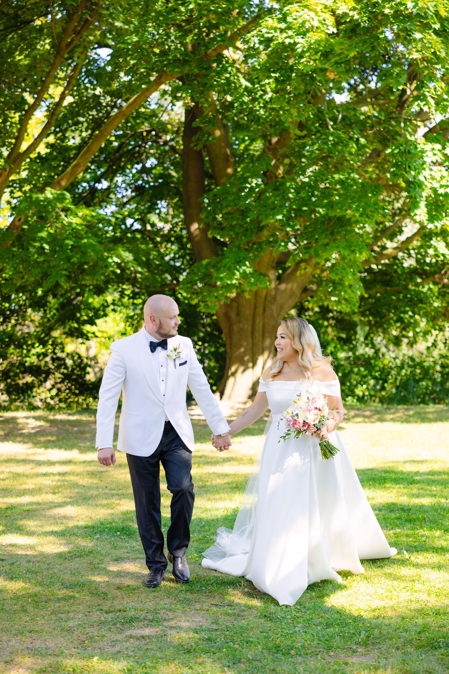 Wedding couple walks hand in hand under green trees on sunny summer day.
