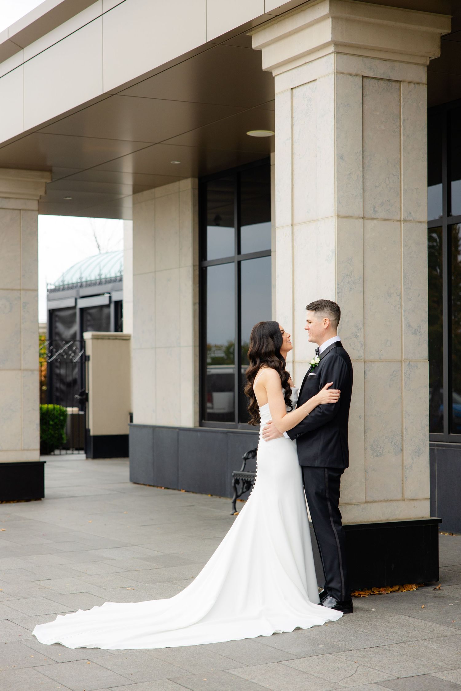 A bride in a flowing white gown and groom in classic tuxedo sharing an intimate moment against stone columns.