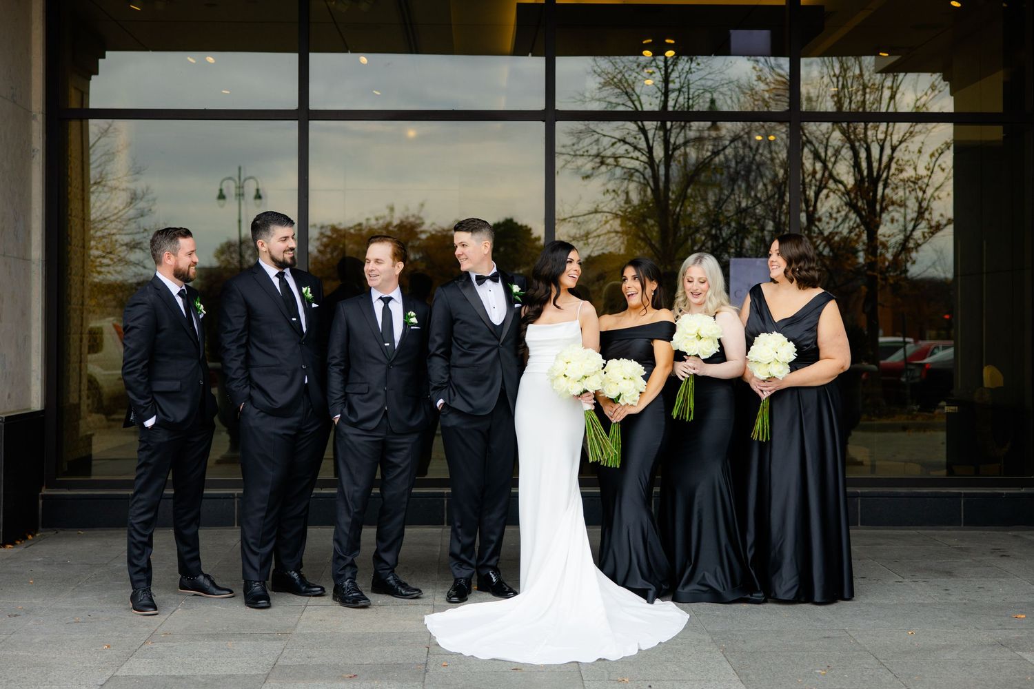 Wedding party with bridesmaids in black dresses and groomsmen in suits pose outdoors.
