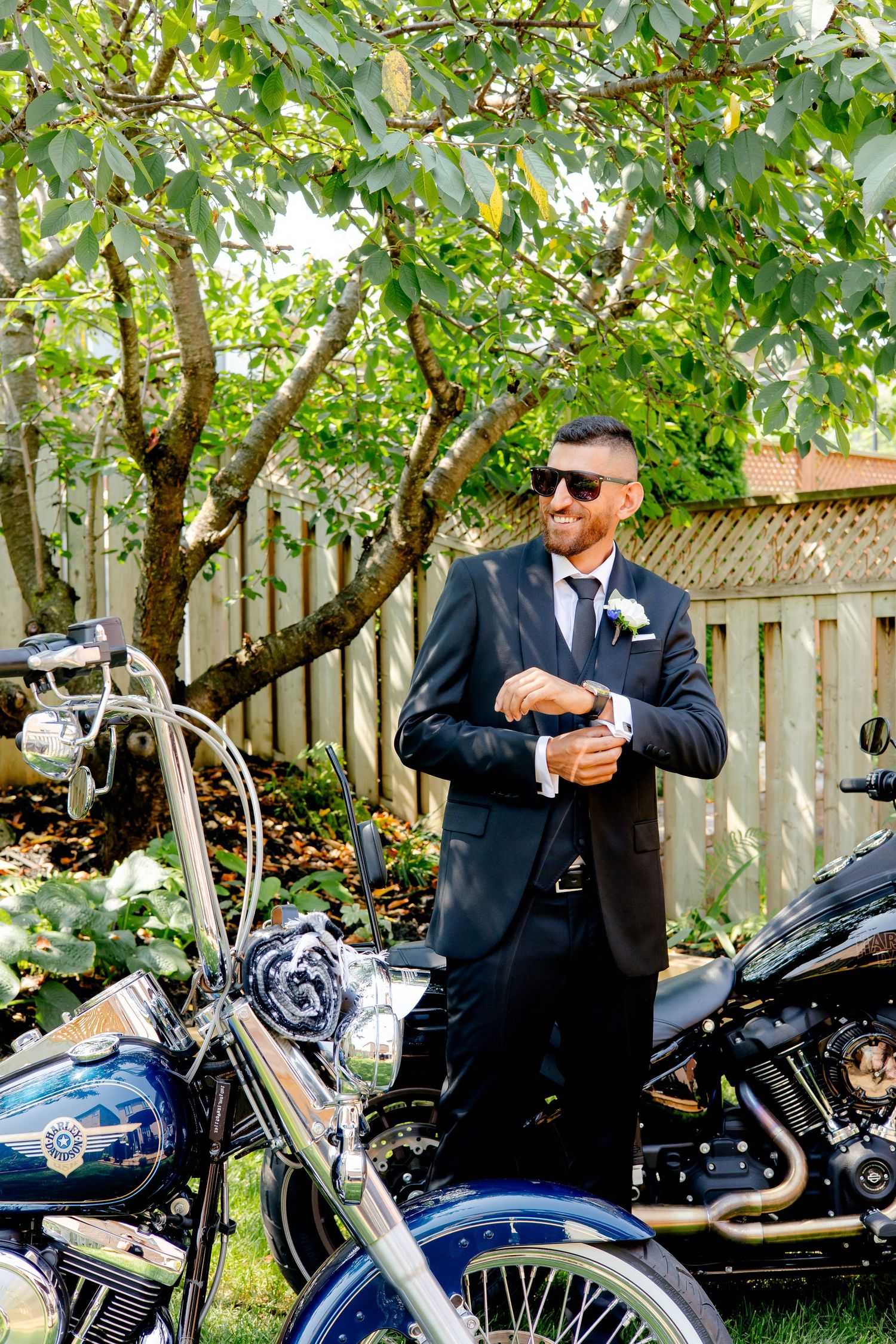 Elegant suited figure checking time next to custom motorcycles under garden trees.