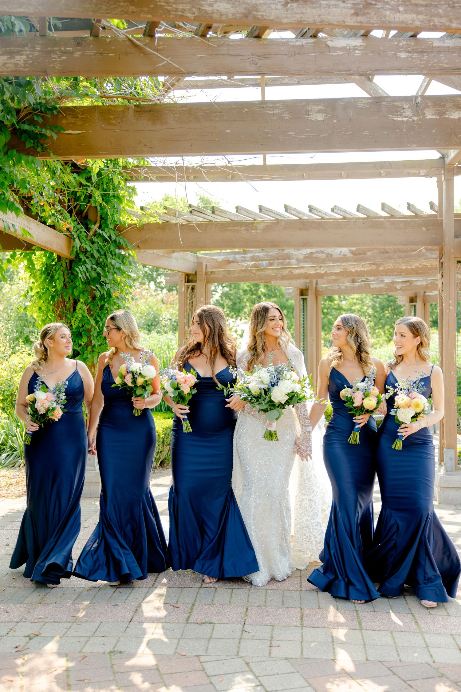 Bridal party in navy and white dresses share candid moment under pergola with climbing vines.