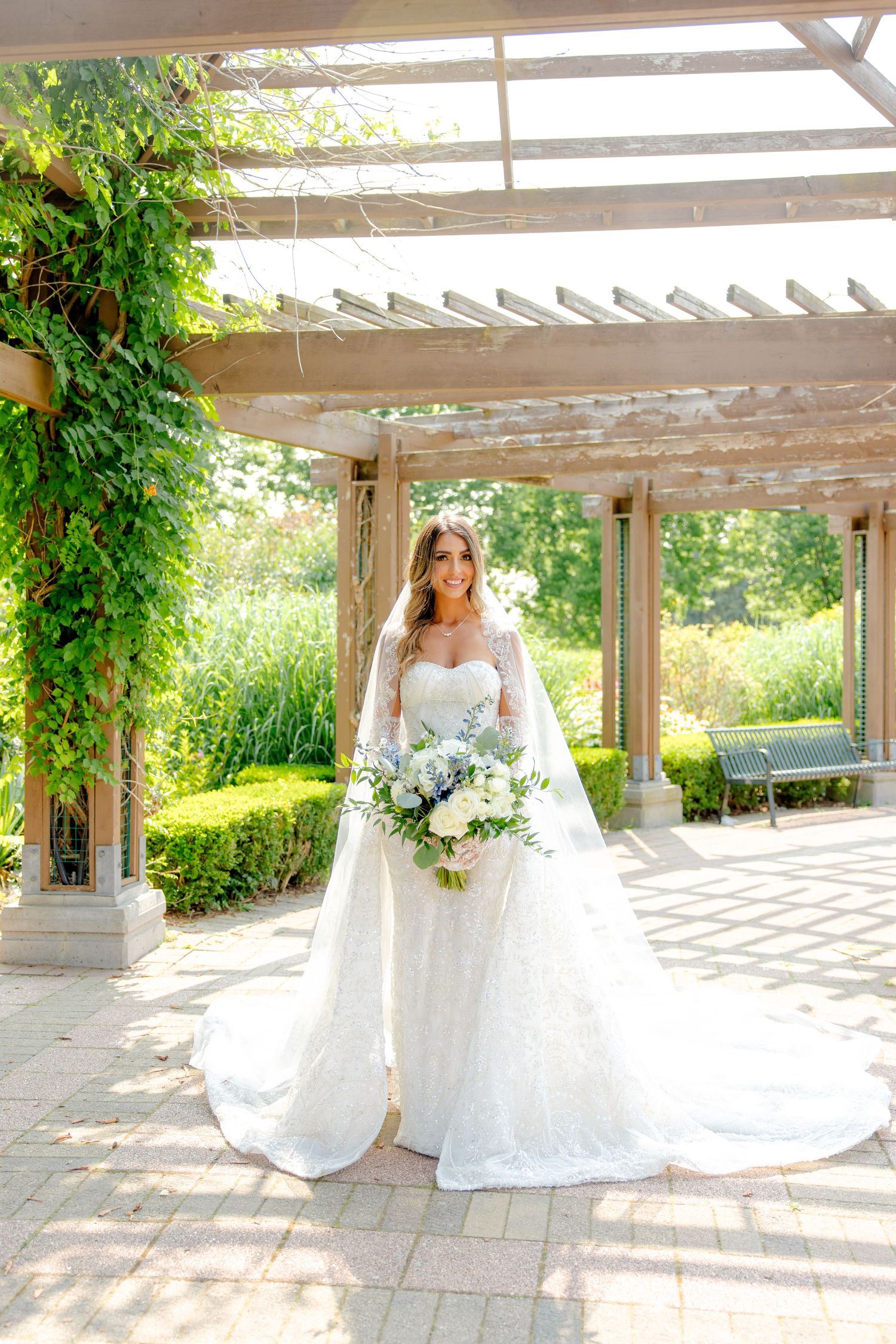 Wedding dress portrait showing elegant lace gown with flowing train under a garden pergola with climbing vines.