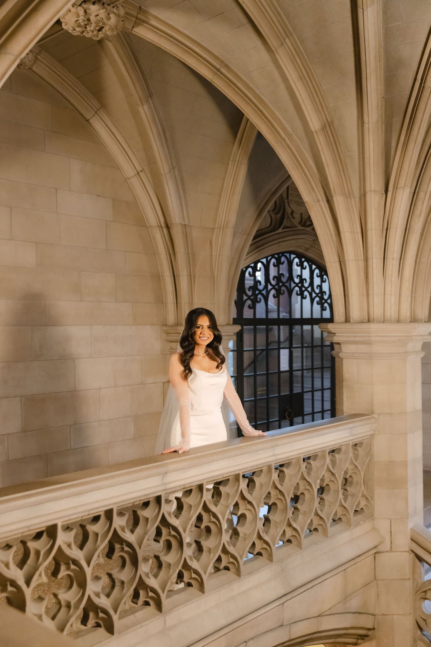 A bride in a white dress stands gracefully on an ornate balcony with gothic stone arches and decorative railings.