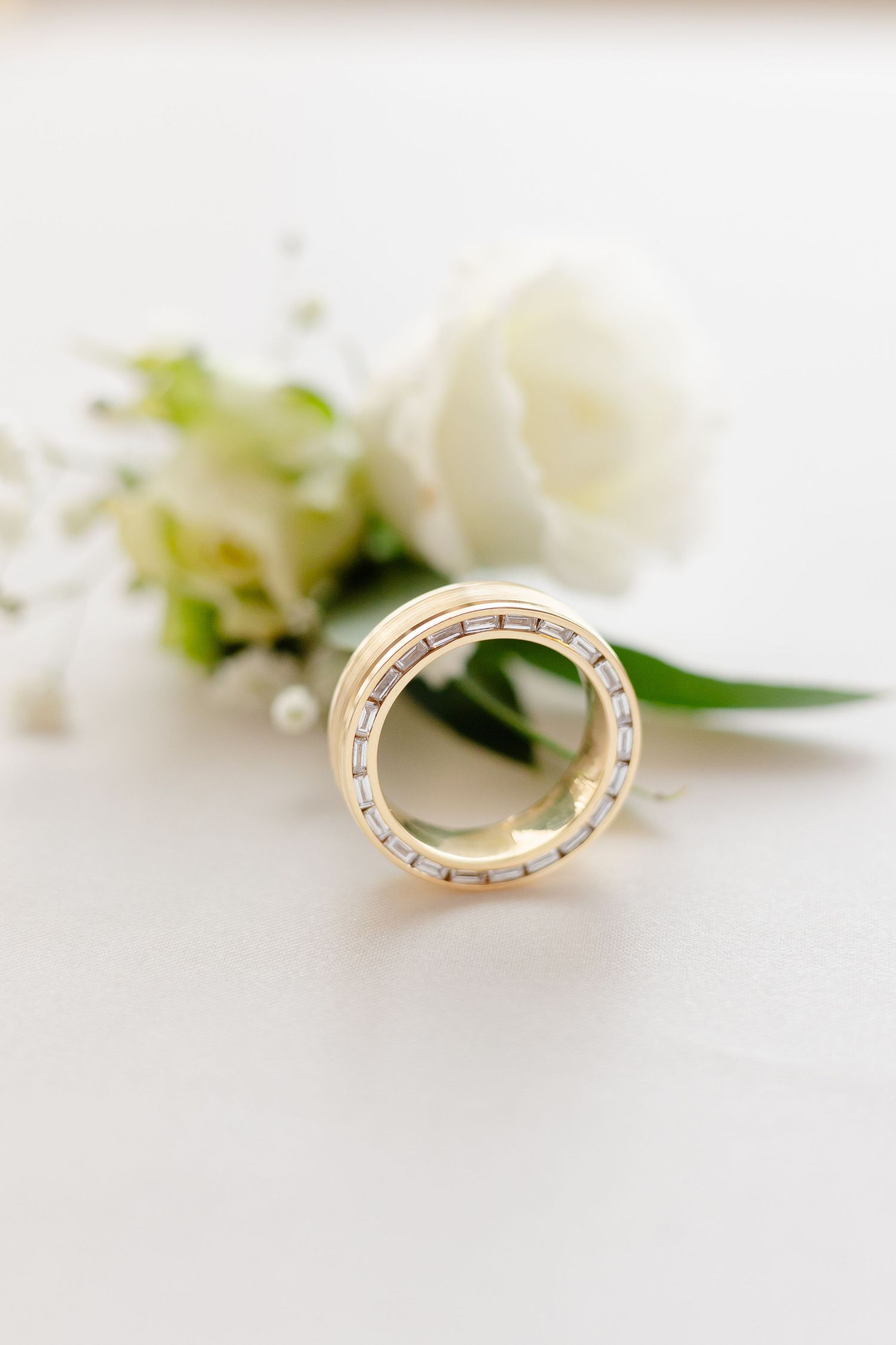 Gold wedding ring with diamonds displayed with a white rose and greenery.
