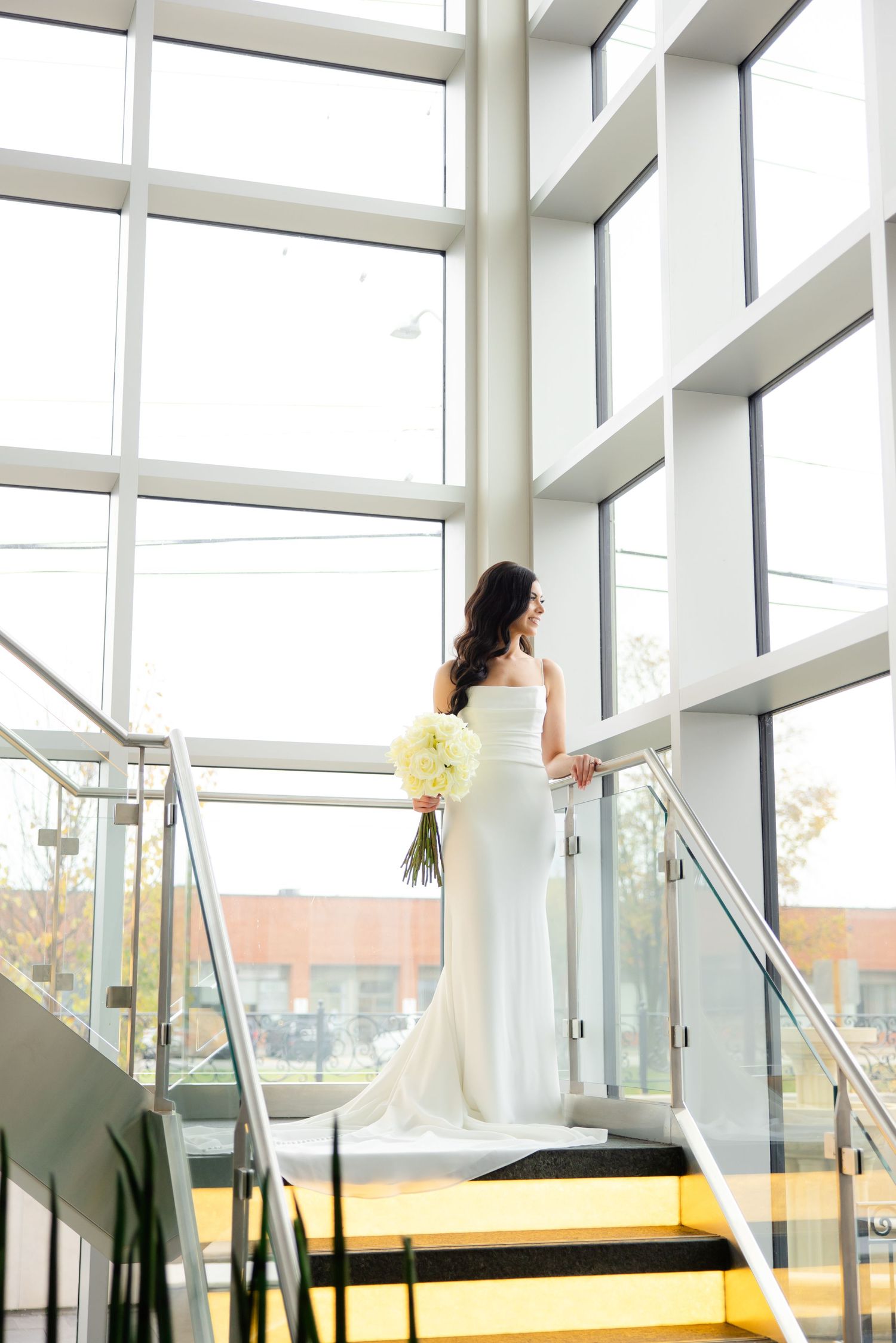 Bride in white gown holds bouquet while standing on modern yellow stairs near floor-to-ceiling windows.