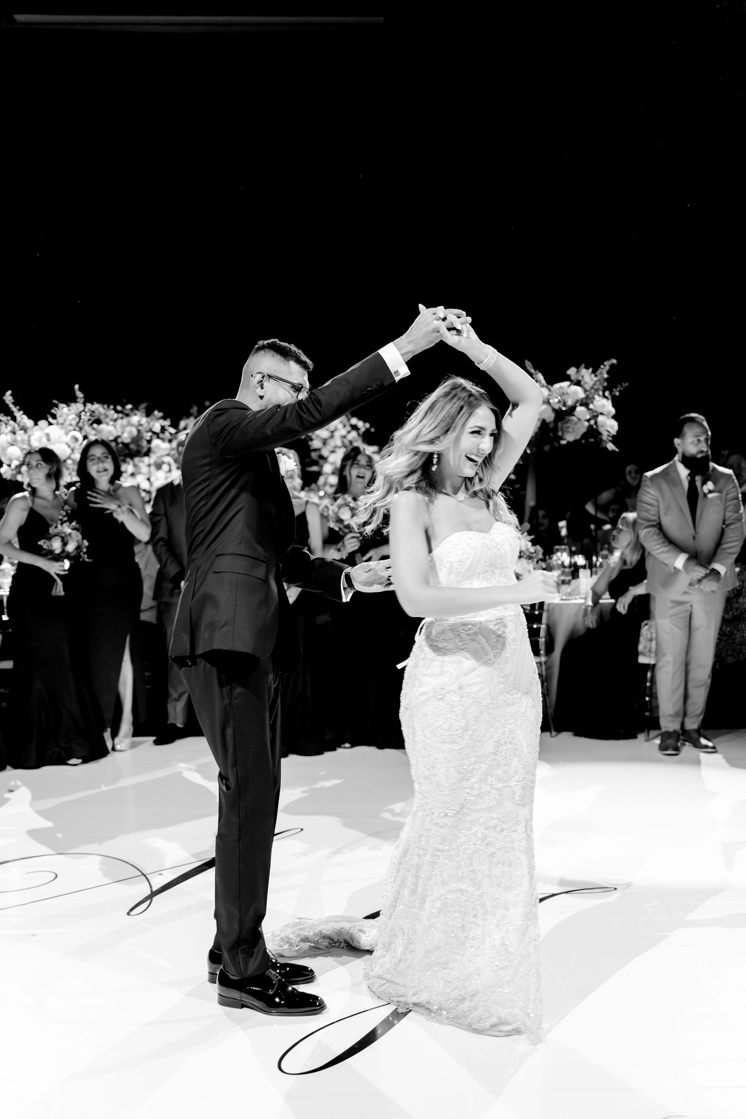 A couple shares their first dance at a wedding reception with a crowd watching in black and white.