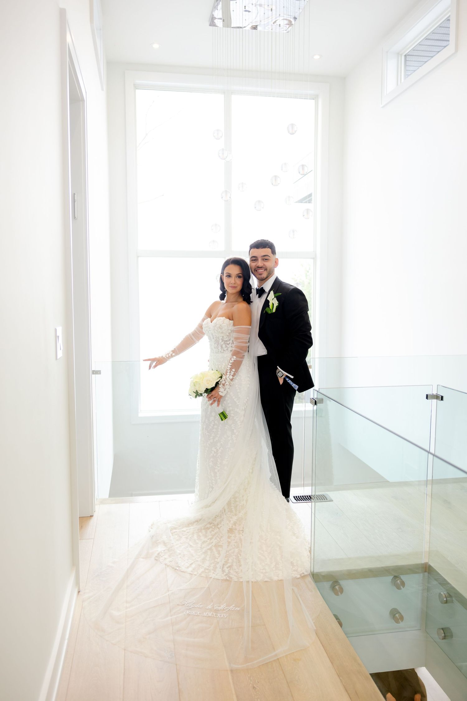 A wedding couple poses on modern glass staircase in bright interior setting.