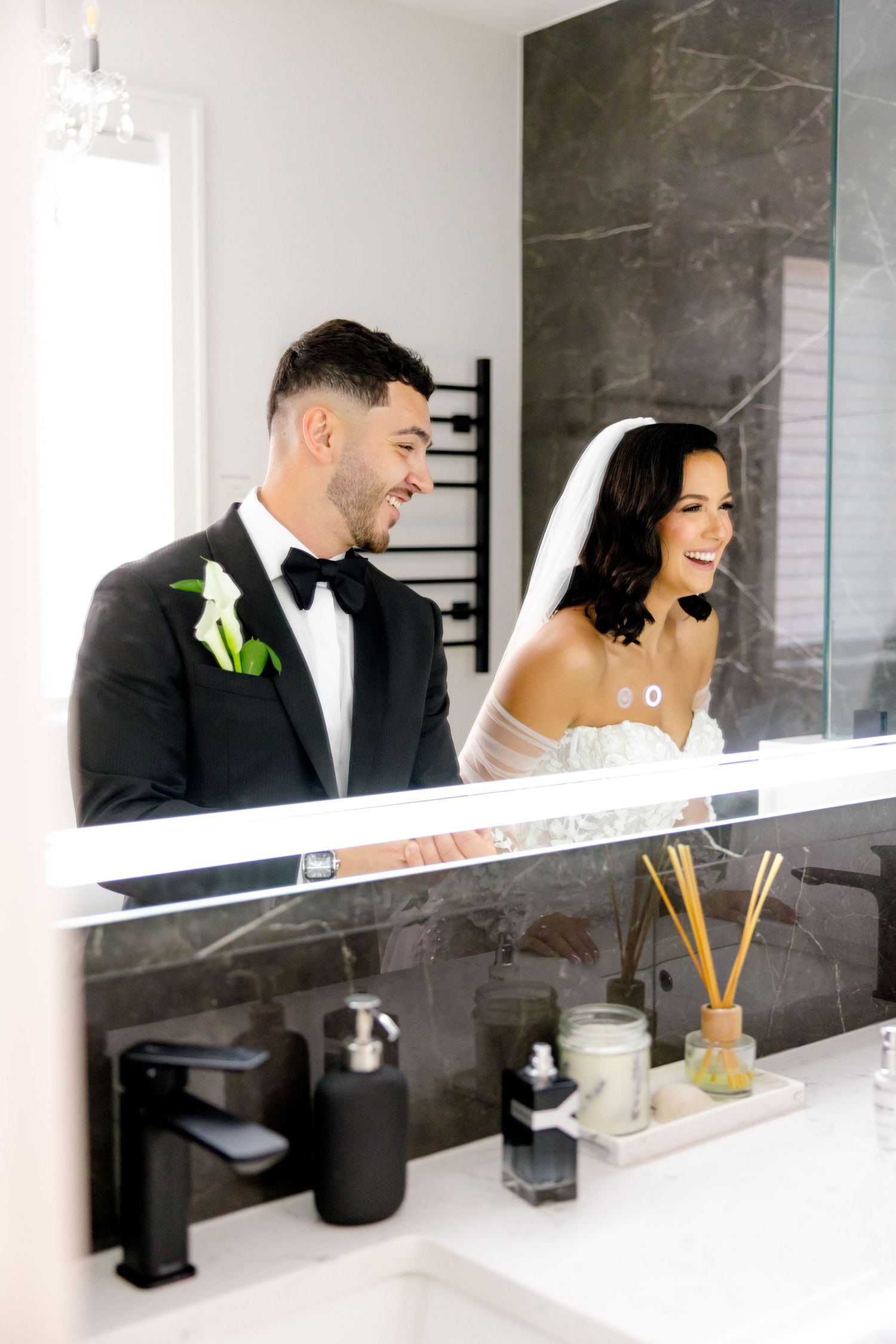 Candid bathroom mirror reflection shows happy newlyweds with modern black fixtures and marble wall.