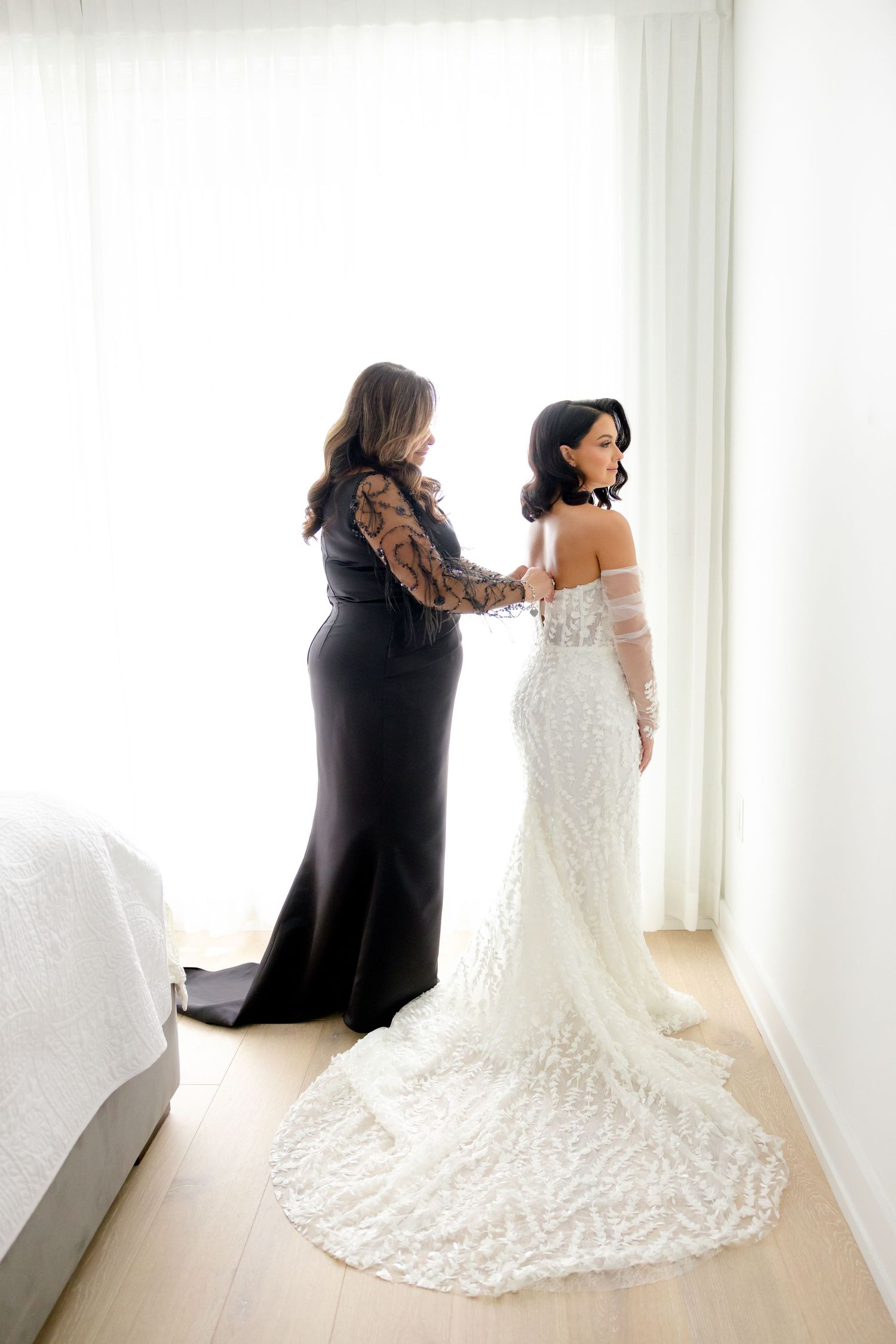 Two people share intimate moment during wedding dress fitting in sunlit room.