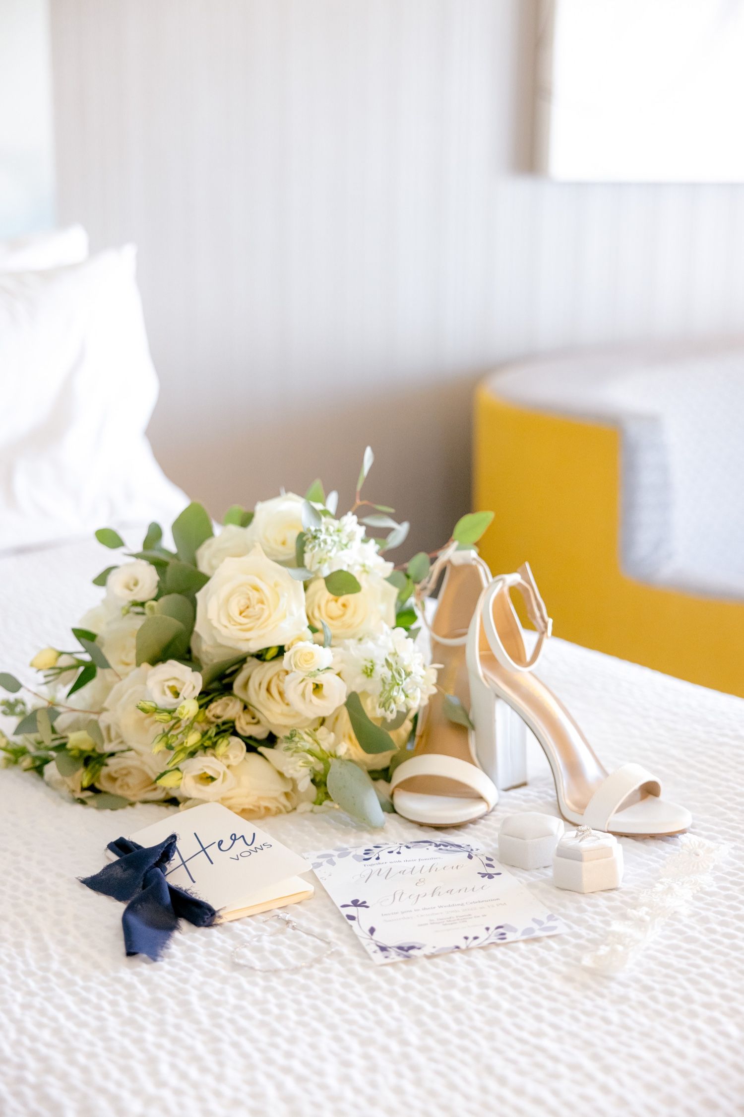 Bridal details with cream roses, white heels, and navy-accented invitations on textured tablecloth.
