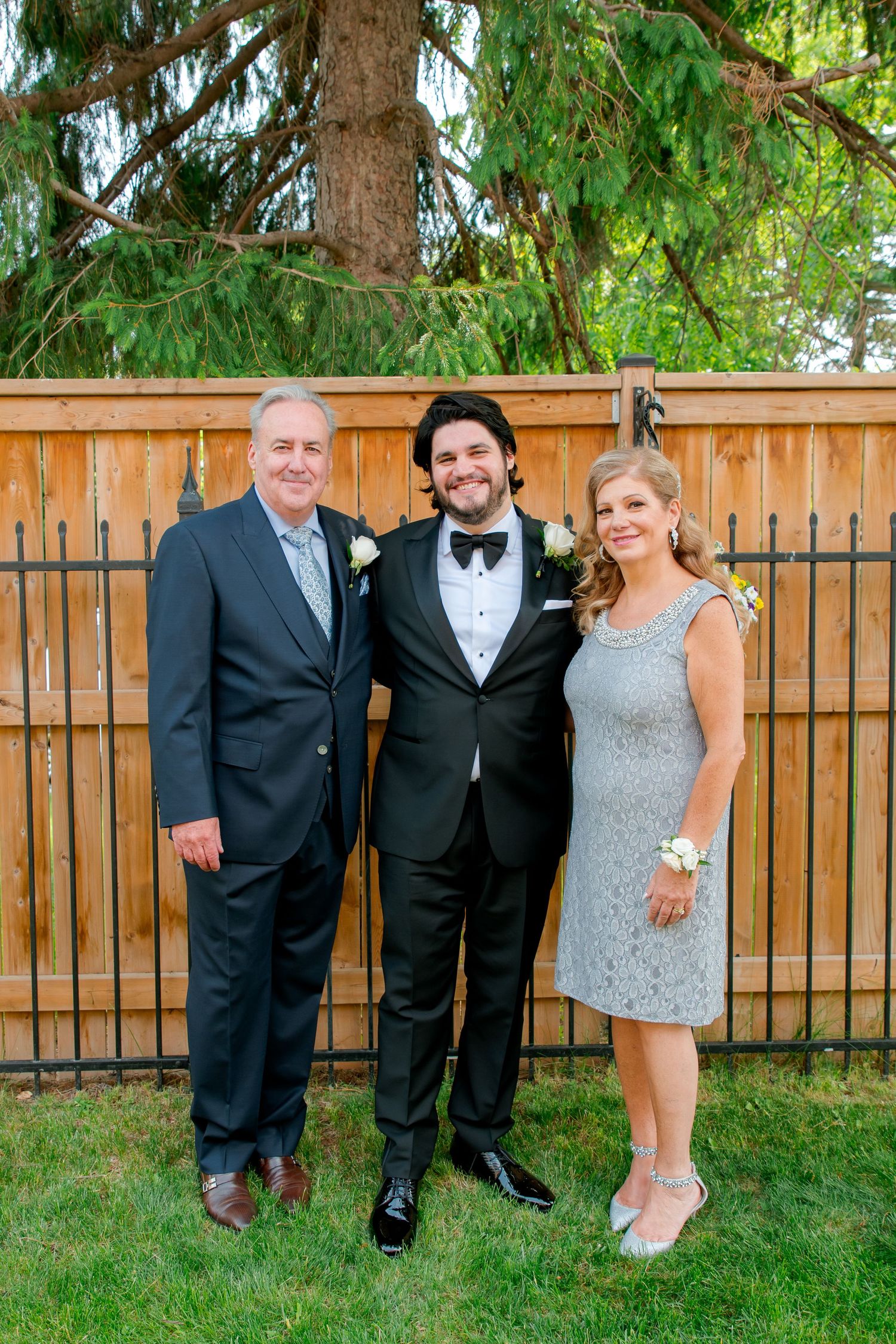 Three people in formal attire pose together against wooden fence with evergreen trees in background.