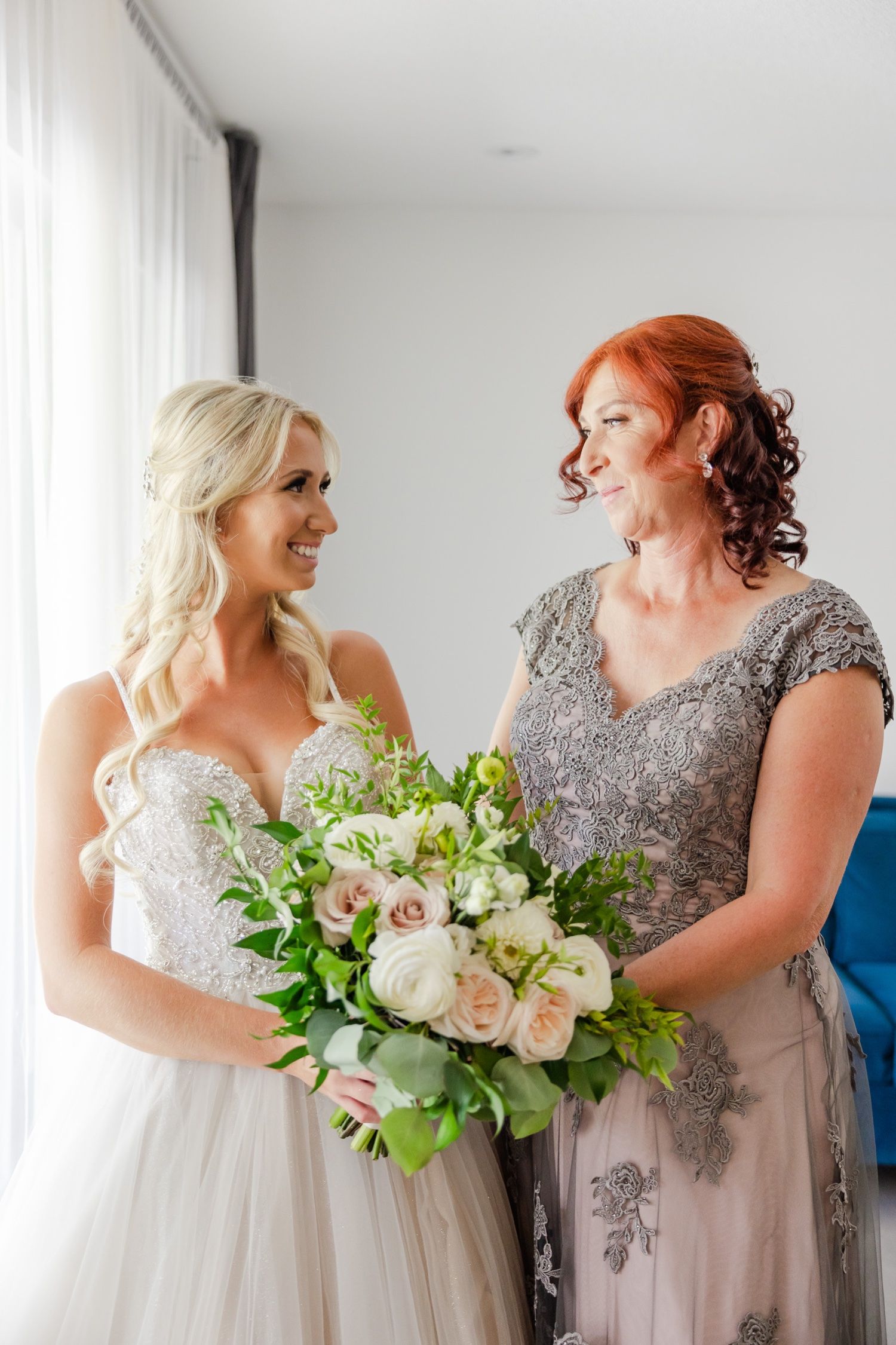 A bride in a white gown and companion in gray lace dress share a joyful moment while holding a lush white bouquet.