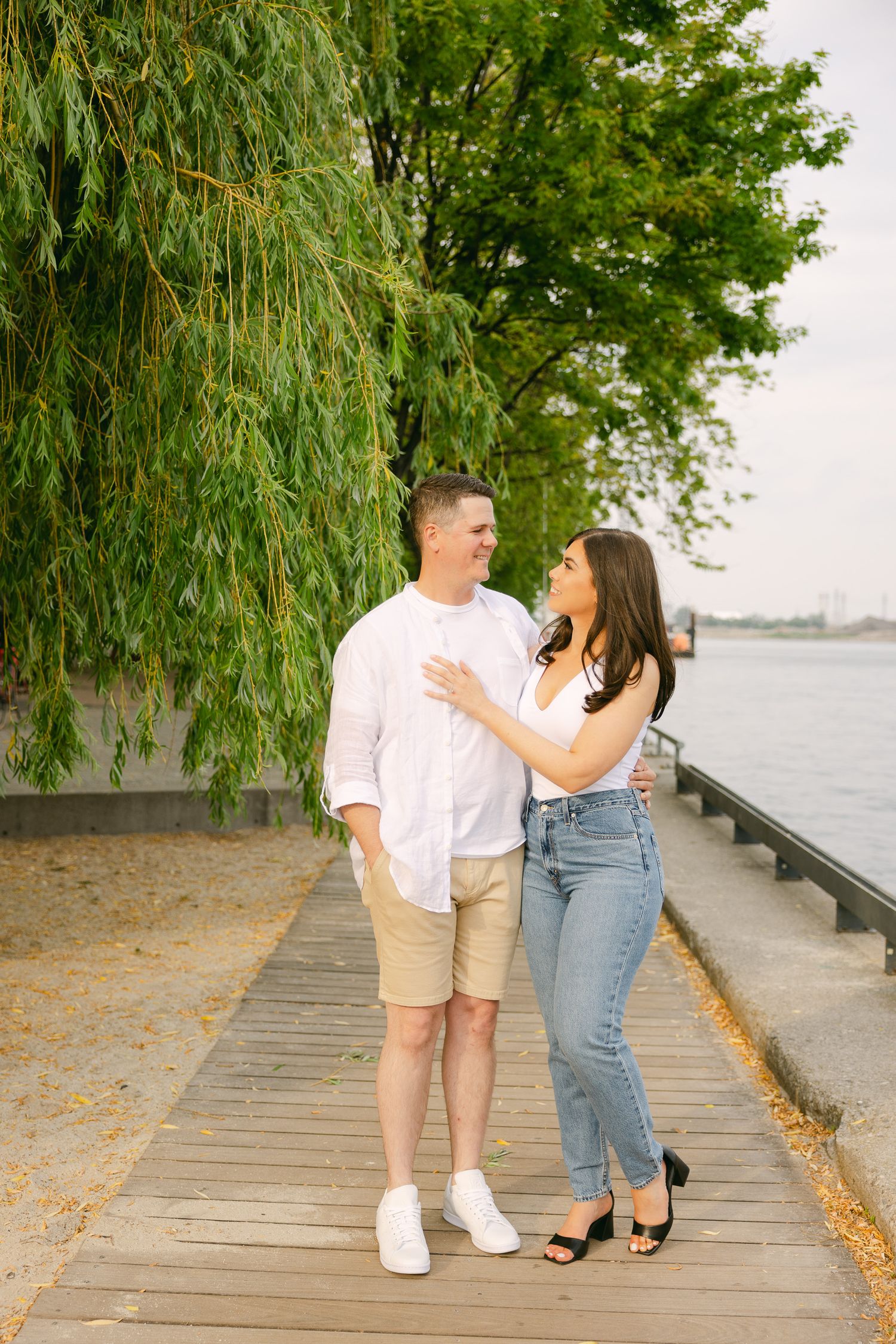 Romantic couple embracing on wooden boardwalk beneath weeping willow tree by waterfront.