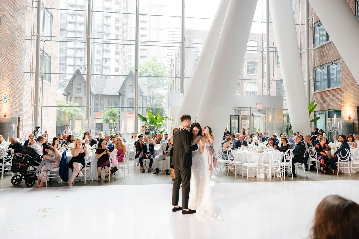 Wedding reception in a modern glass-walled venue with guests seated at round tables.