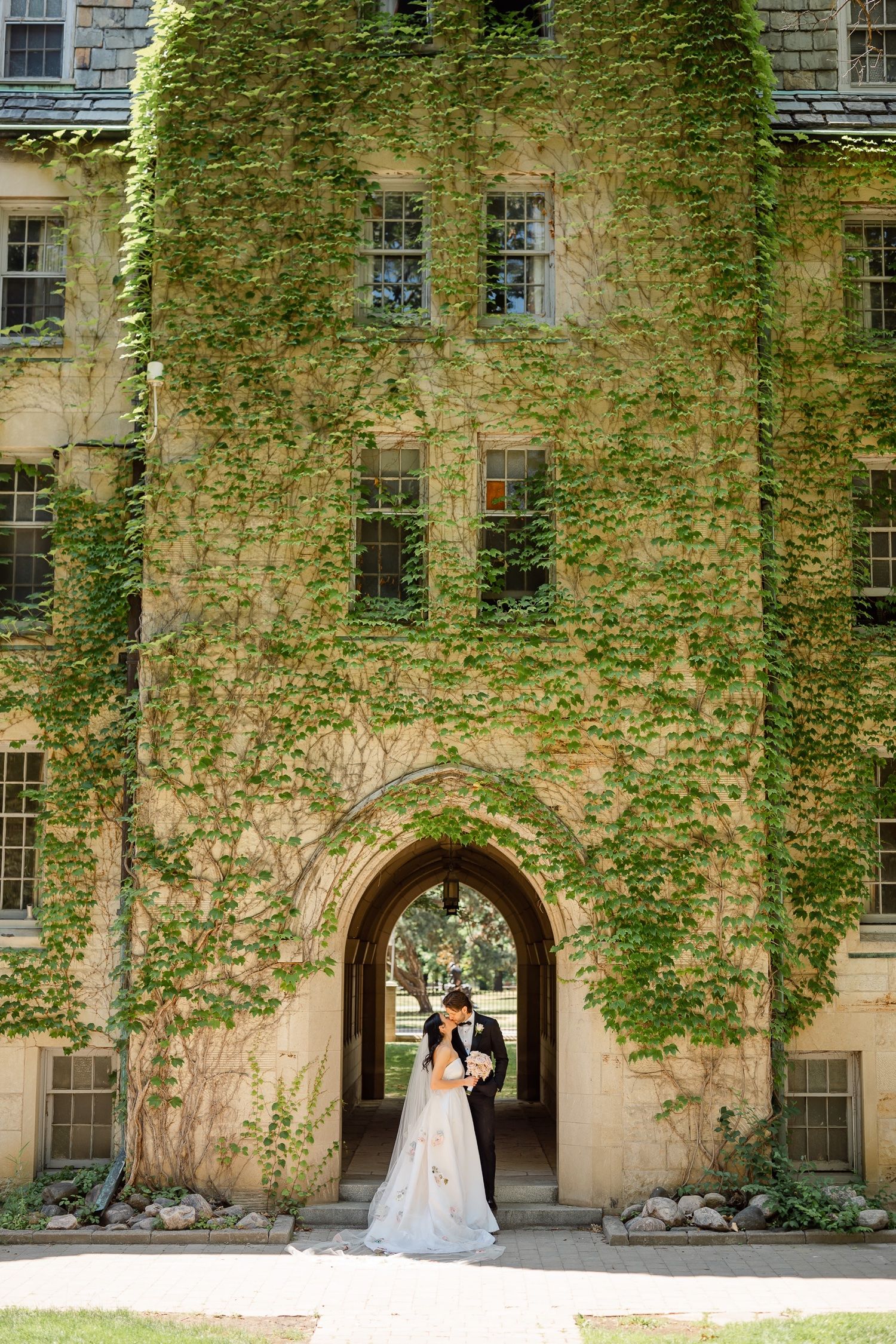 Sunlit wedding portrait at entrance of ivy-covered stone building with gothic architecture.