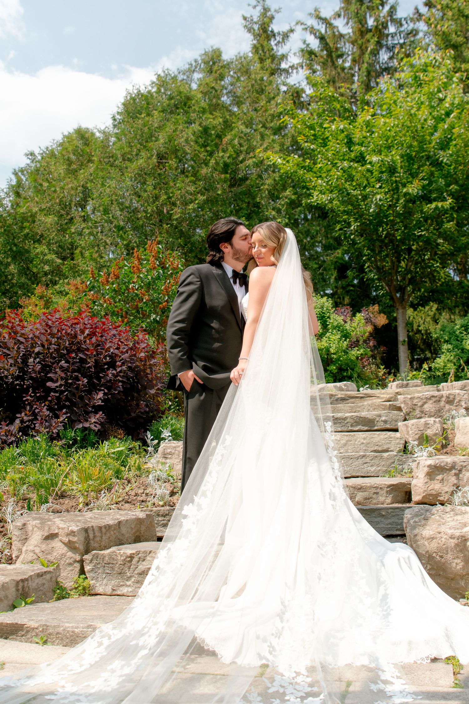 Wedding couple embraces on stone garden steps with dramatic cathedral veil trailing behind.