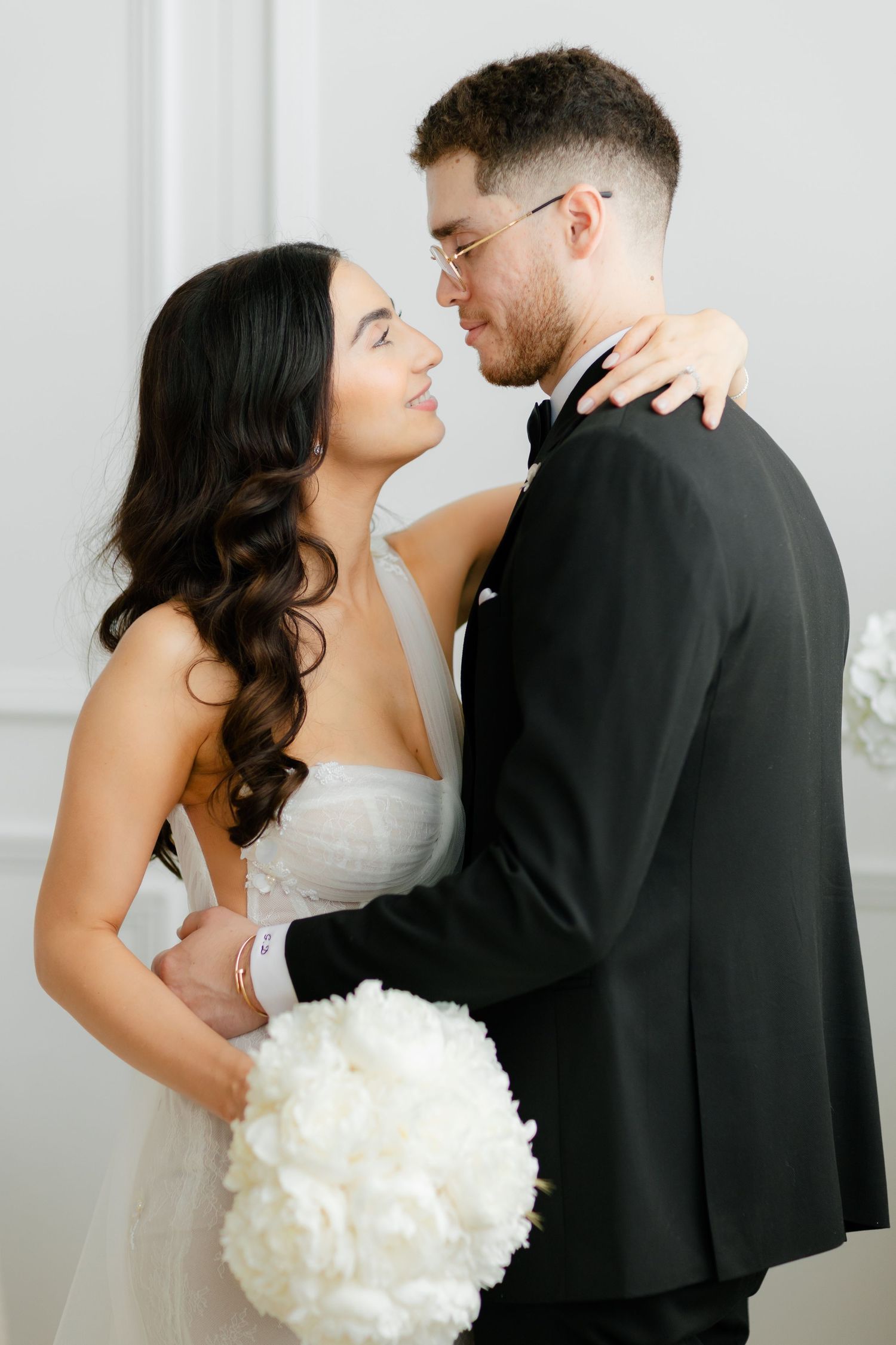 Intimate wedding portrait shows couple embracing against white wall.