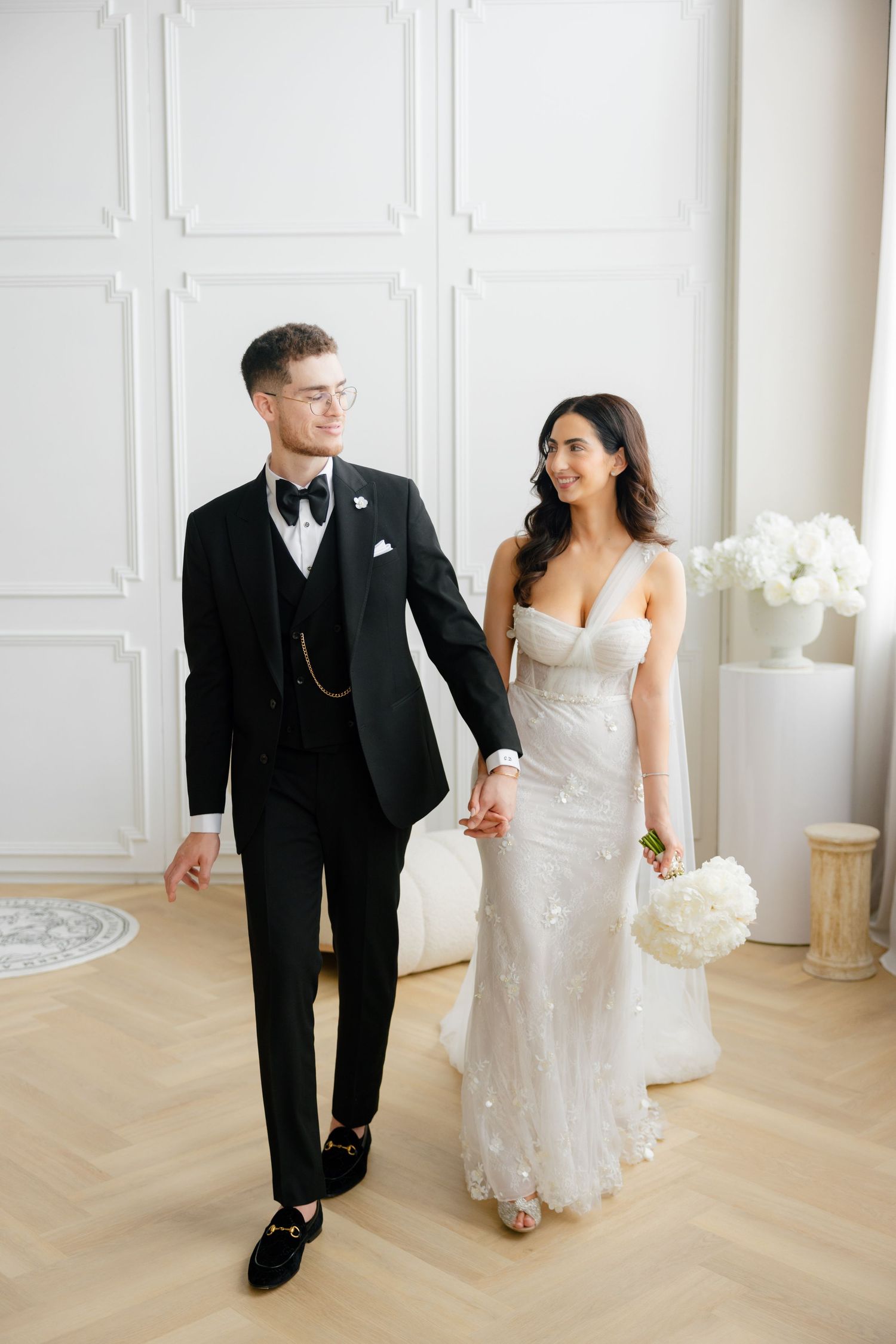 A couple in elegant wedding attire walks together in a bright white room with classic wainscoting and floral decor.