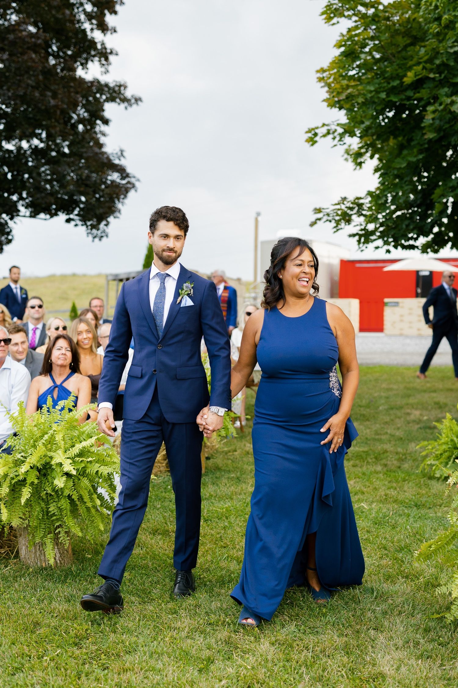 Wedding processional with attendants in navy blue formal attire walking across a grassy lawn.