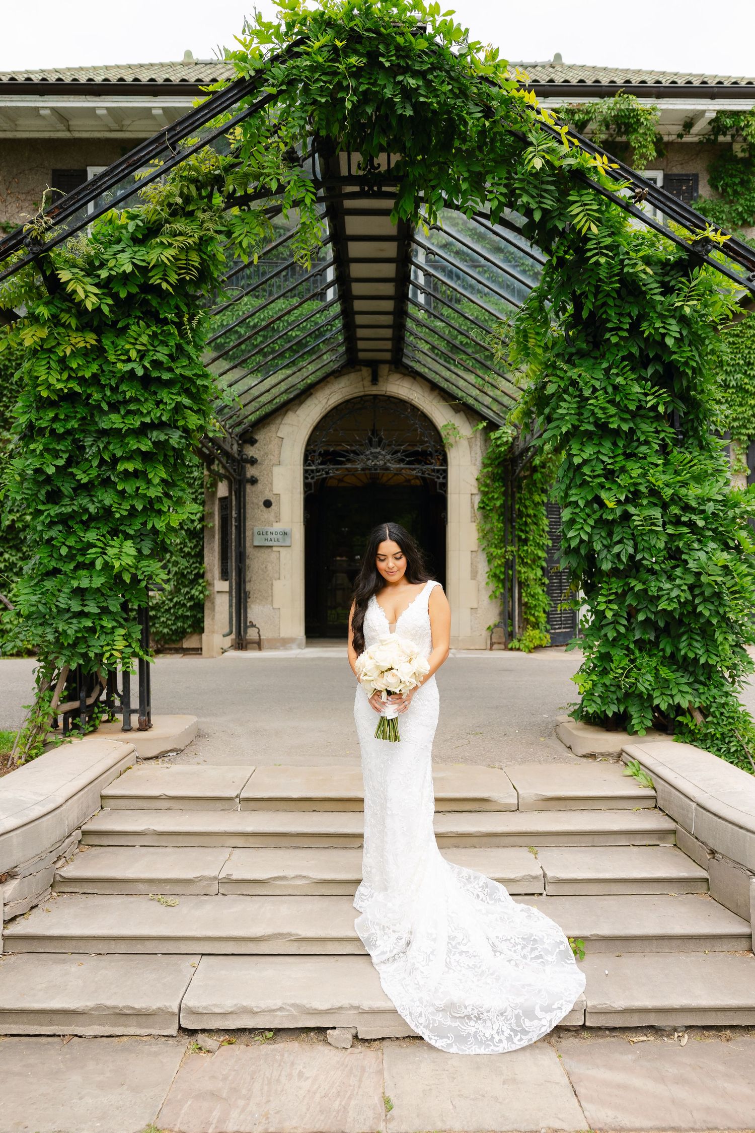 Bride in fitted lace gown with cathedral train stands on stone steps under ivy-covered glass conservatory.