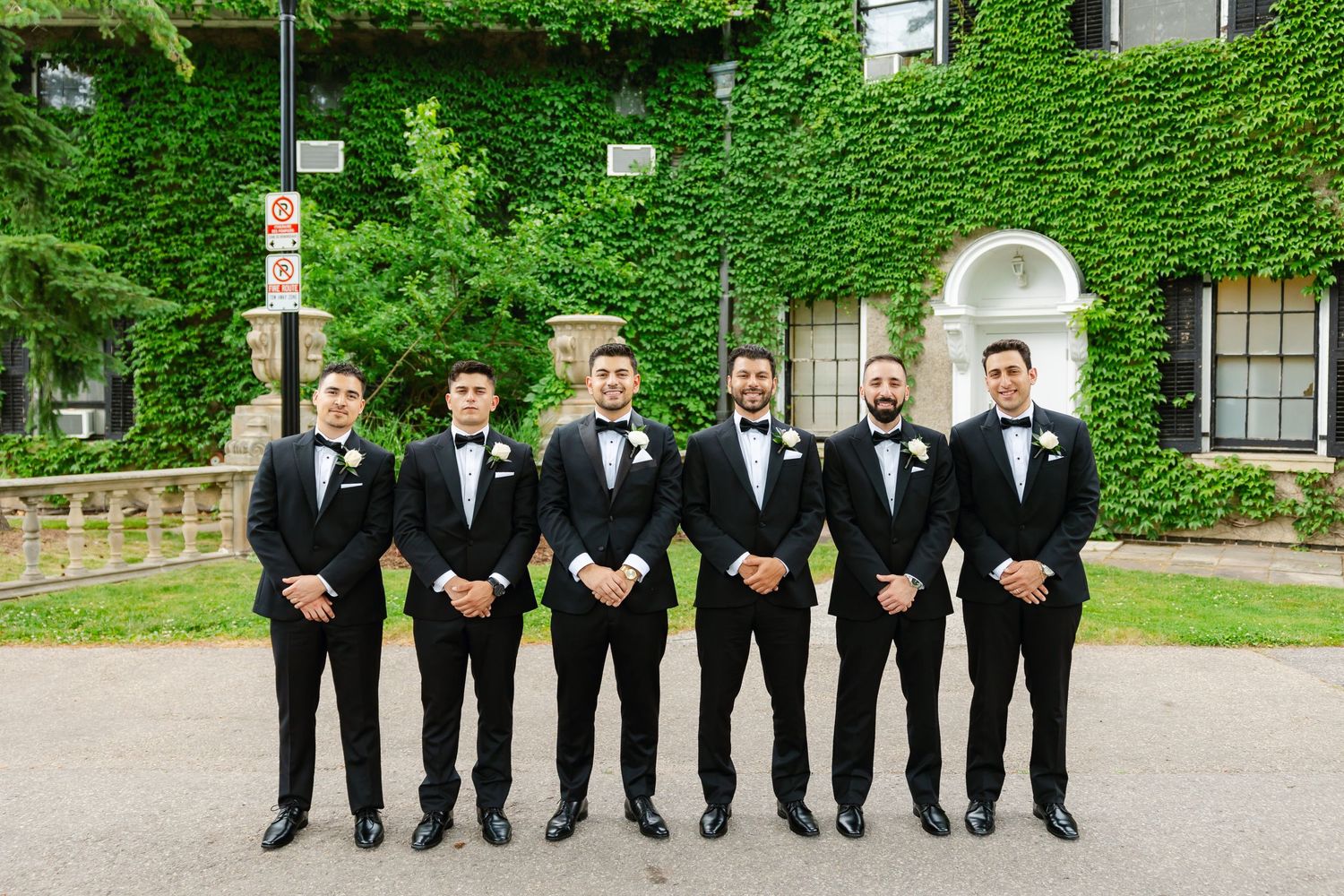 Groomsmen lined up in black tuxedos pose formally against an ivy-covered building with arched doorway.