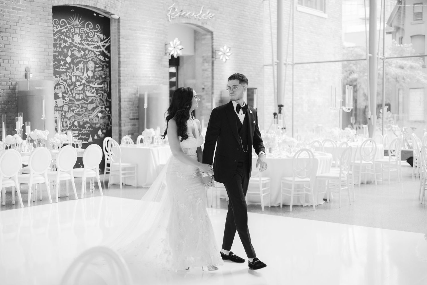 Black and white photo of newlyweds walking through their reception space.