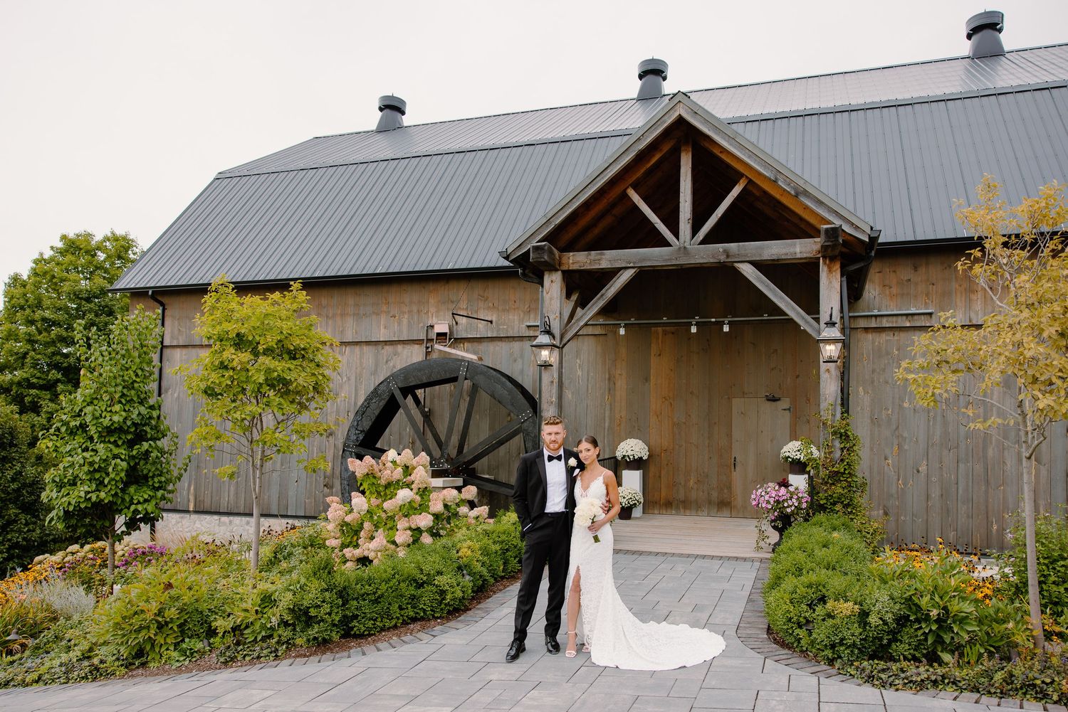 A couple poses outside a modern barn venue with grey metal roof and landscaped entrance.