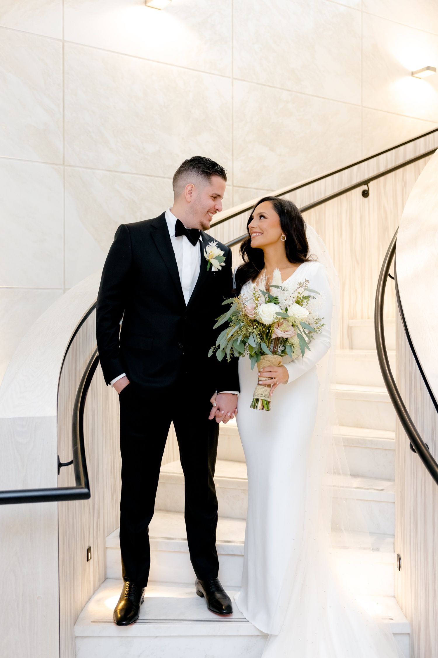 A bride in sleek white dress holds a bouquet while standing with groom in tuxedo on elegant white staircase.