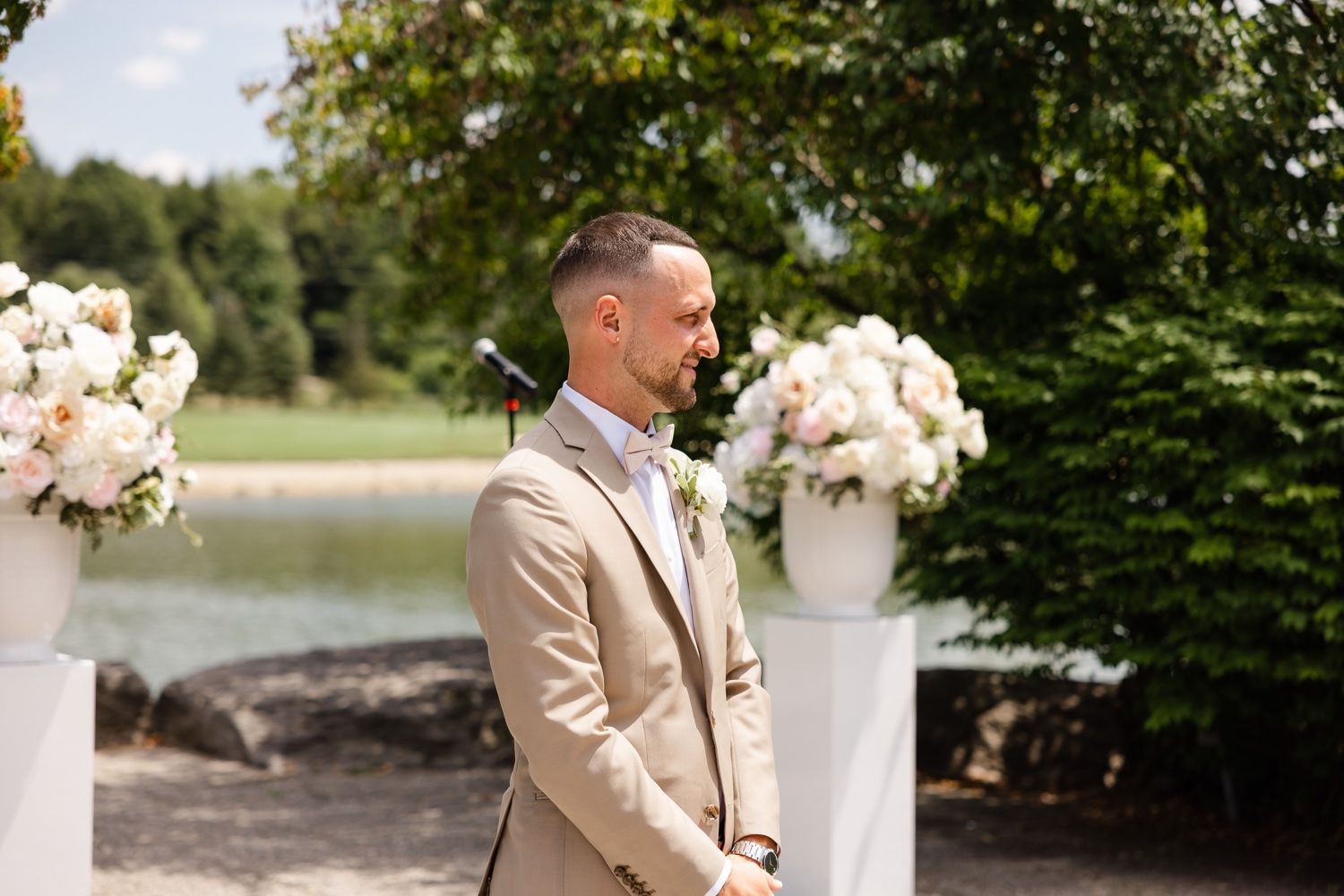 A groom in a beige suit stands at an outdoor wedding ceremony setup with white floral arrangements and lake views.