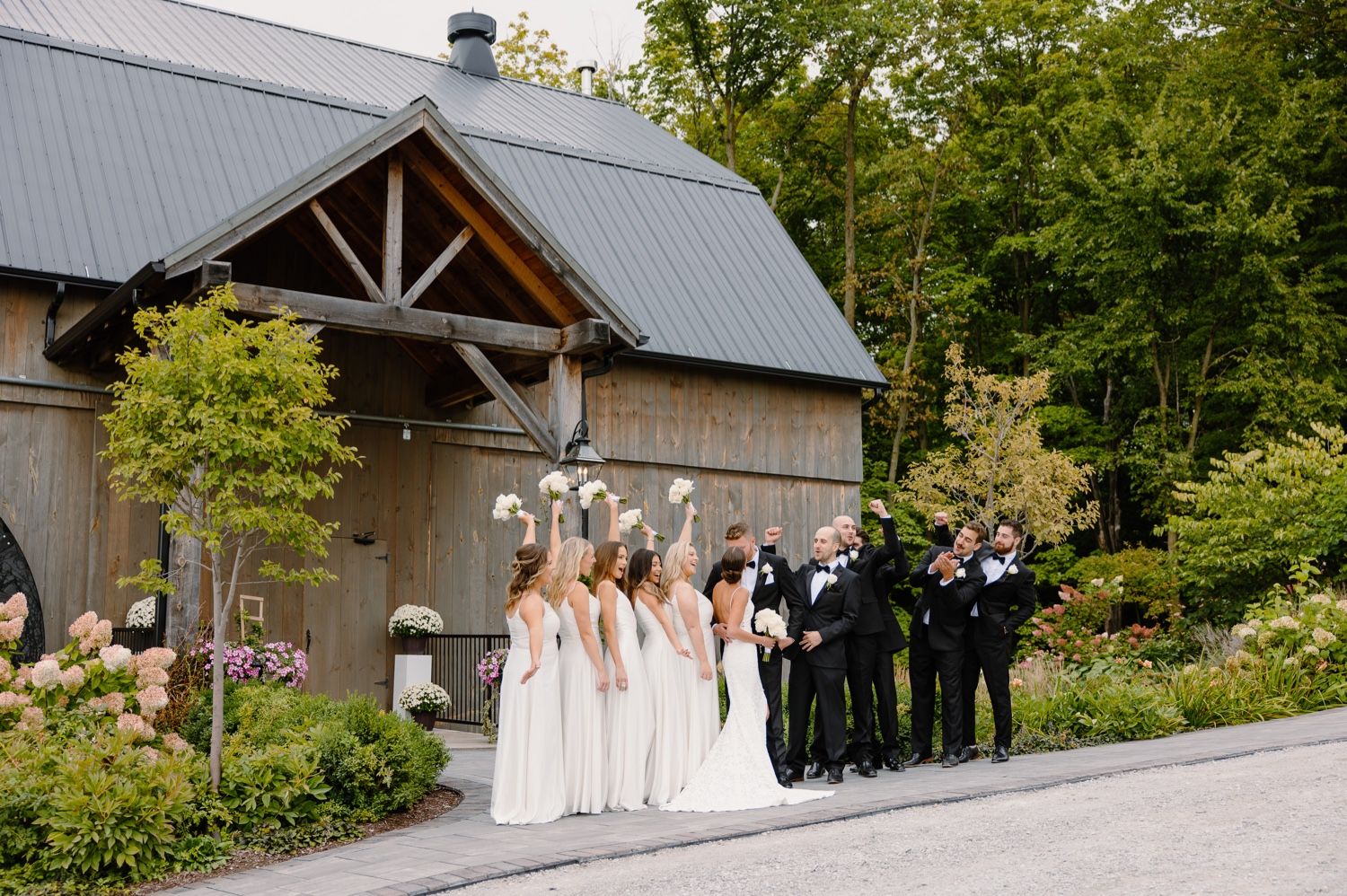 Wedding party in formal attire poses along driveway near rustic barn and gardens.