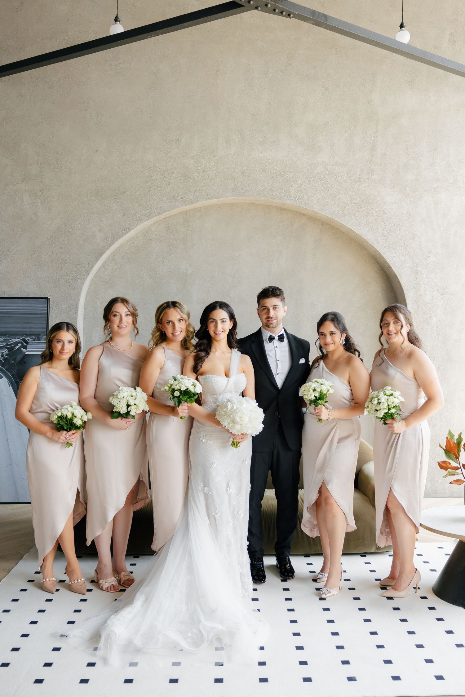 Bridal party in blush dresses stands together under arched wall with geometric floor tiles.
