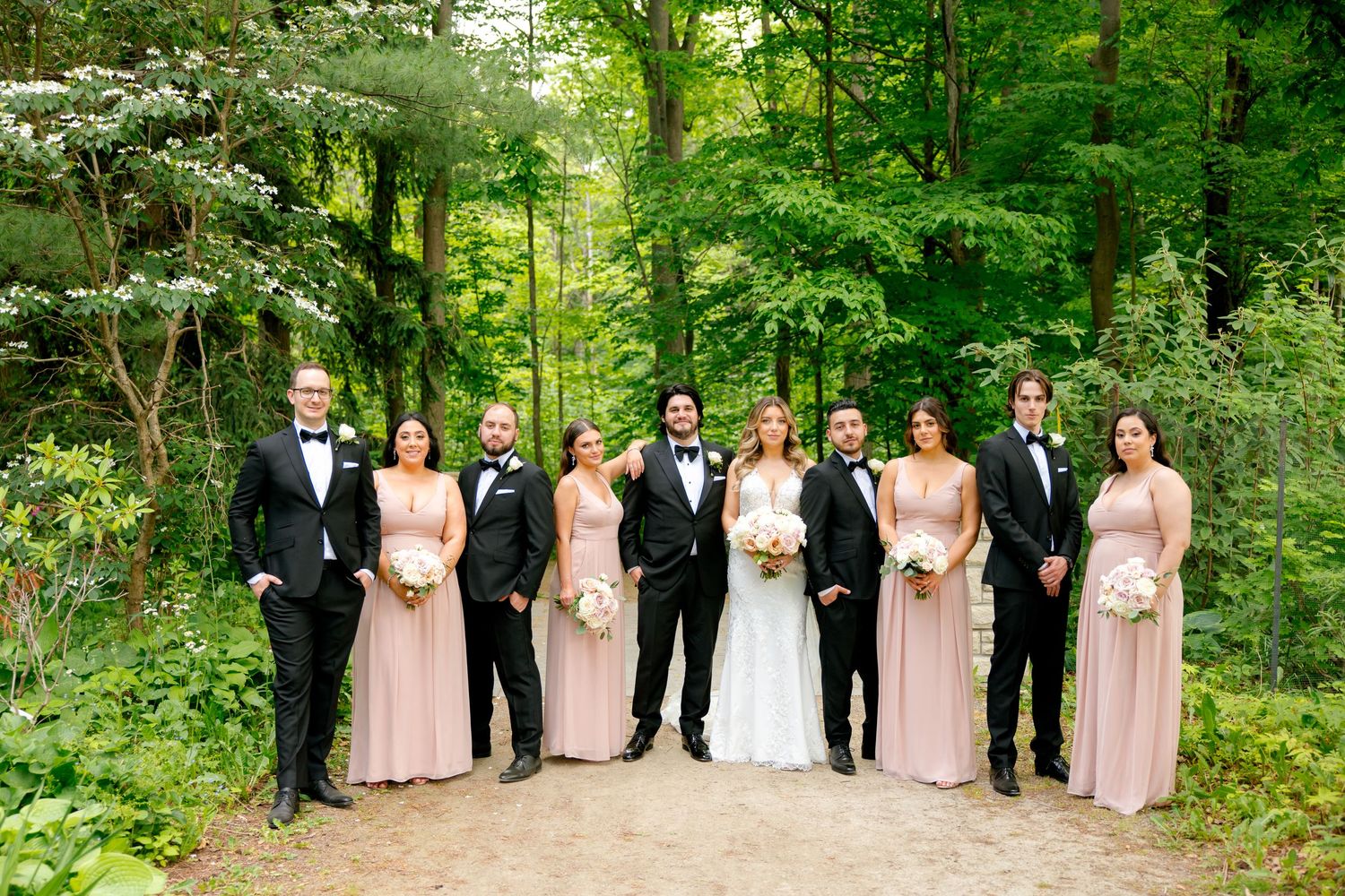 Wedding party group photo on wooded path with pink bridesmaid dresses and classic tuxedos.