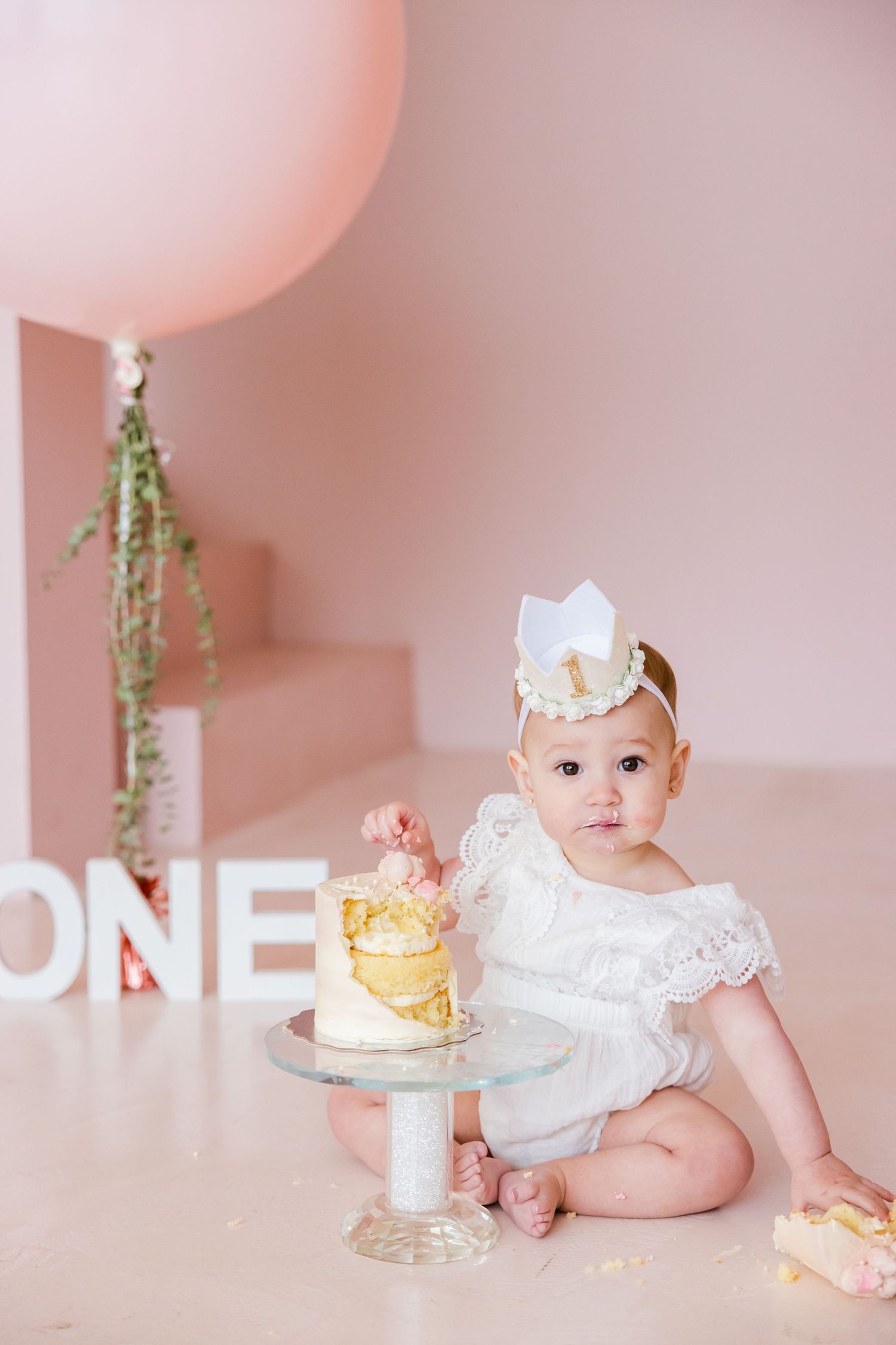 First birthday cake smash setup with white decor, pink balloon, and gold accents on a pink background.