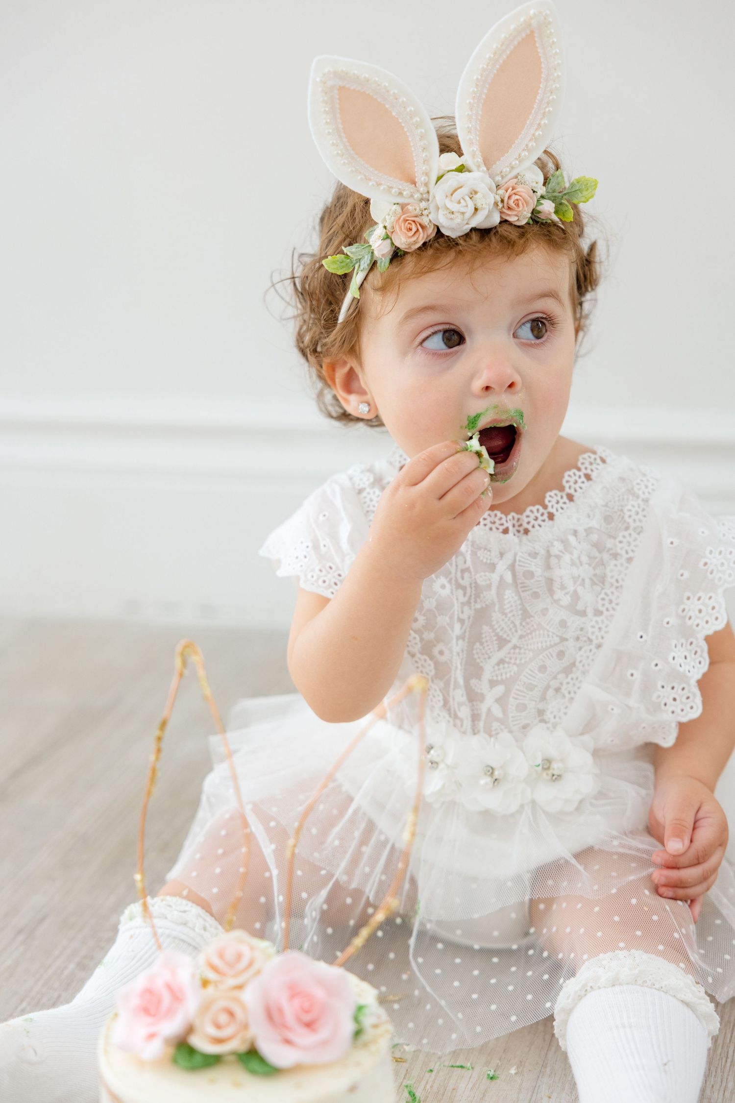 A baby in a white lace dress and bunny ear crown looks excited while eating cake at a party.