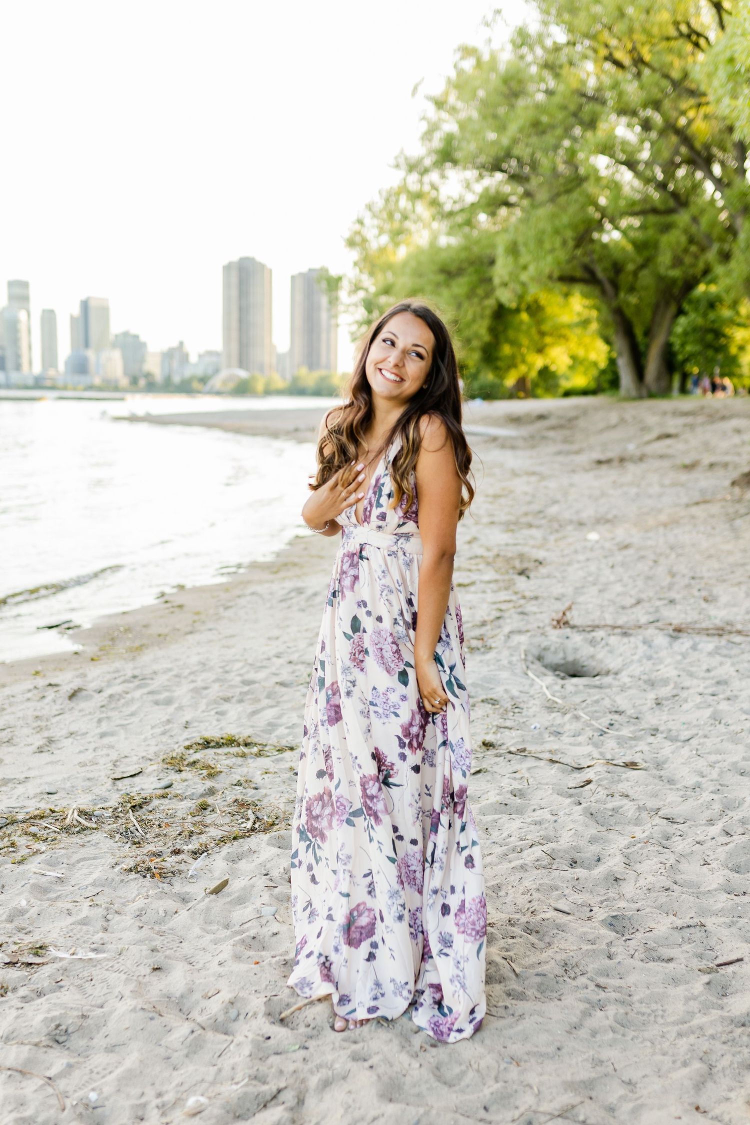 A woman in a floral maxi dress smiles brightly on a beach with cityscape behind her.