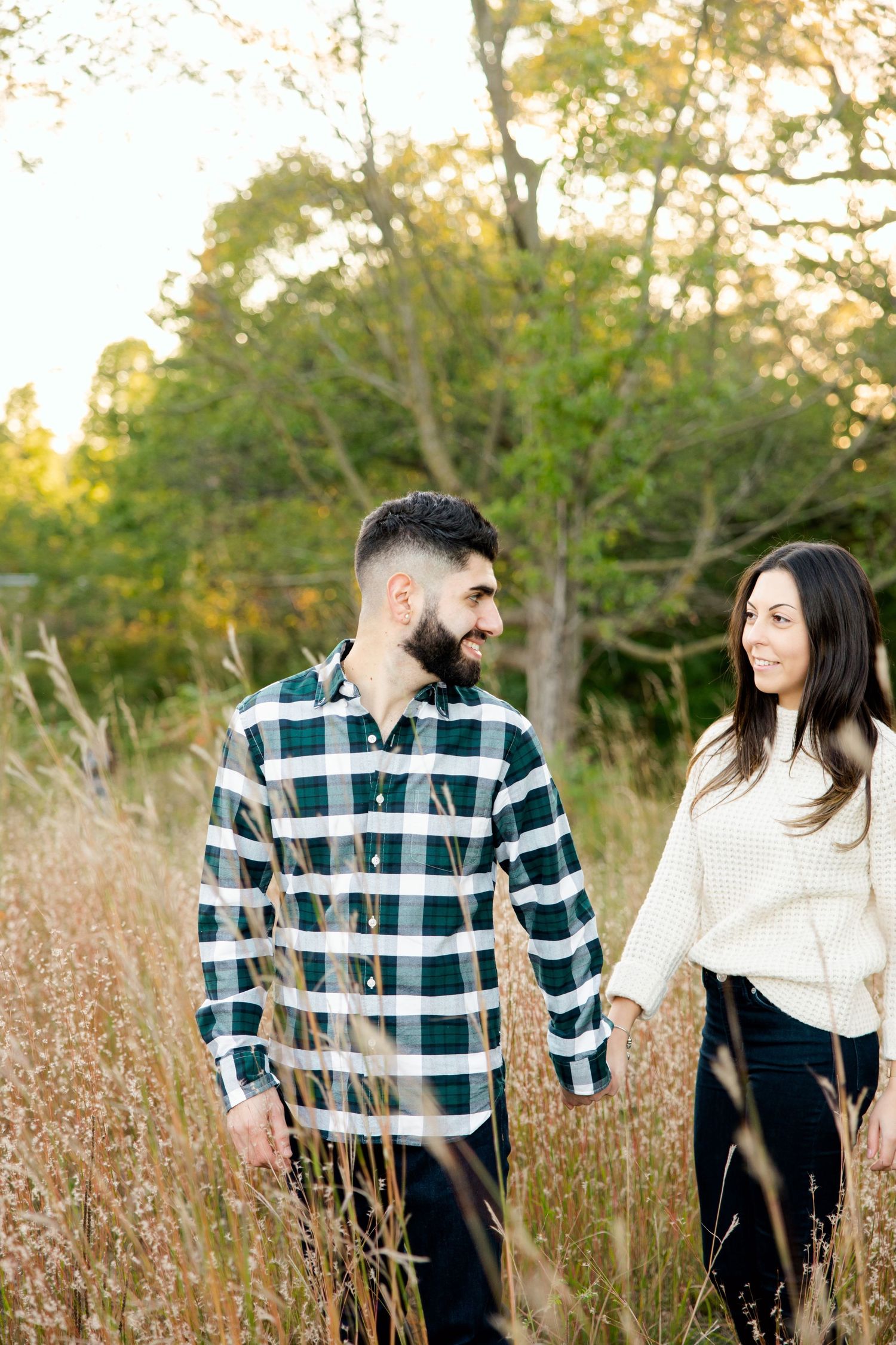Couple walking through tall autumn grass wearing plaid shirt and cream sweater.