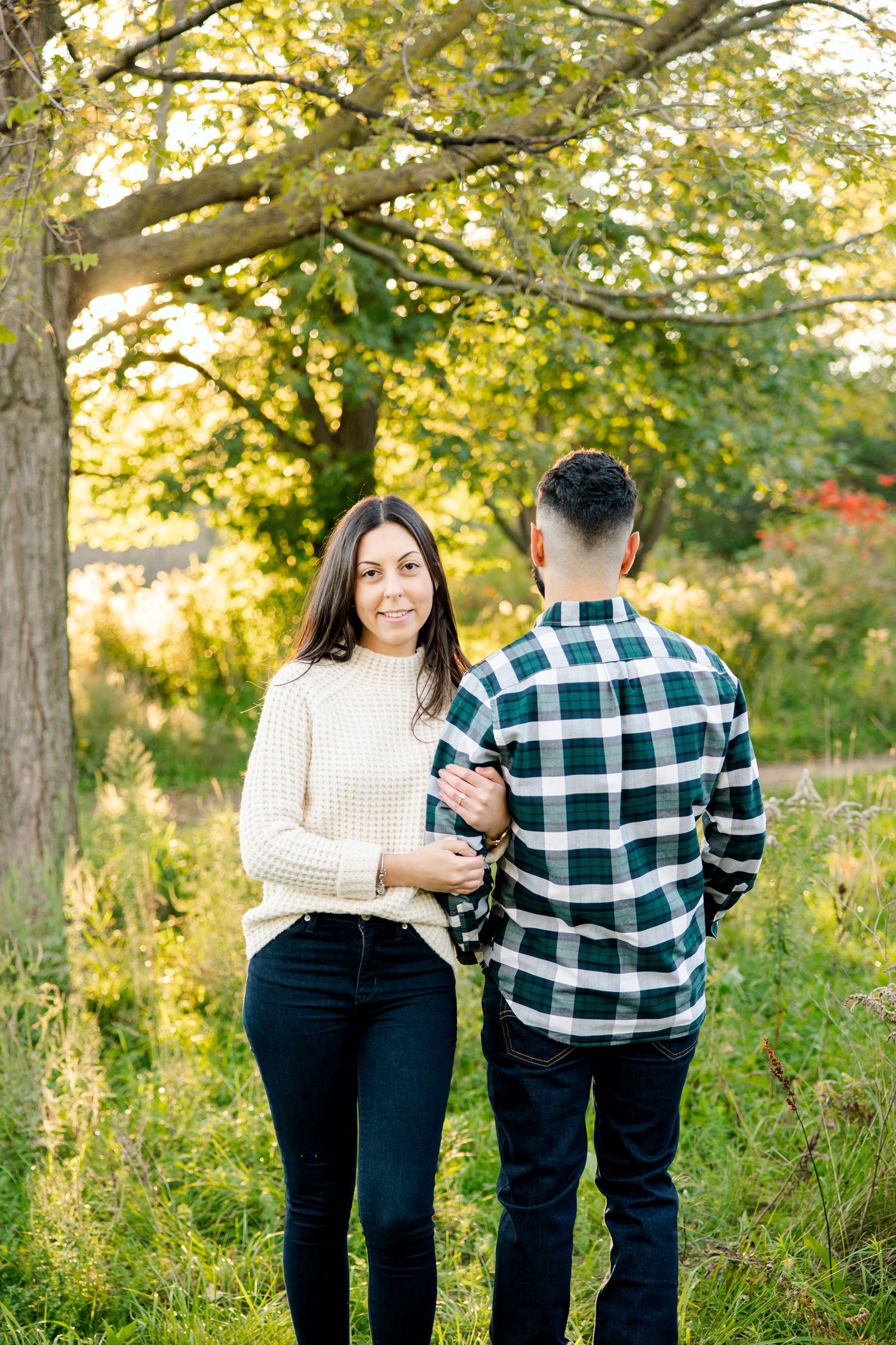 Couple in casual attire standing together under tree with golden fall foliage.