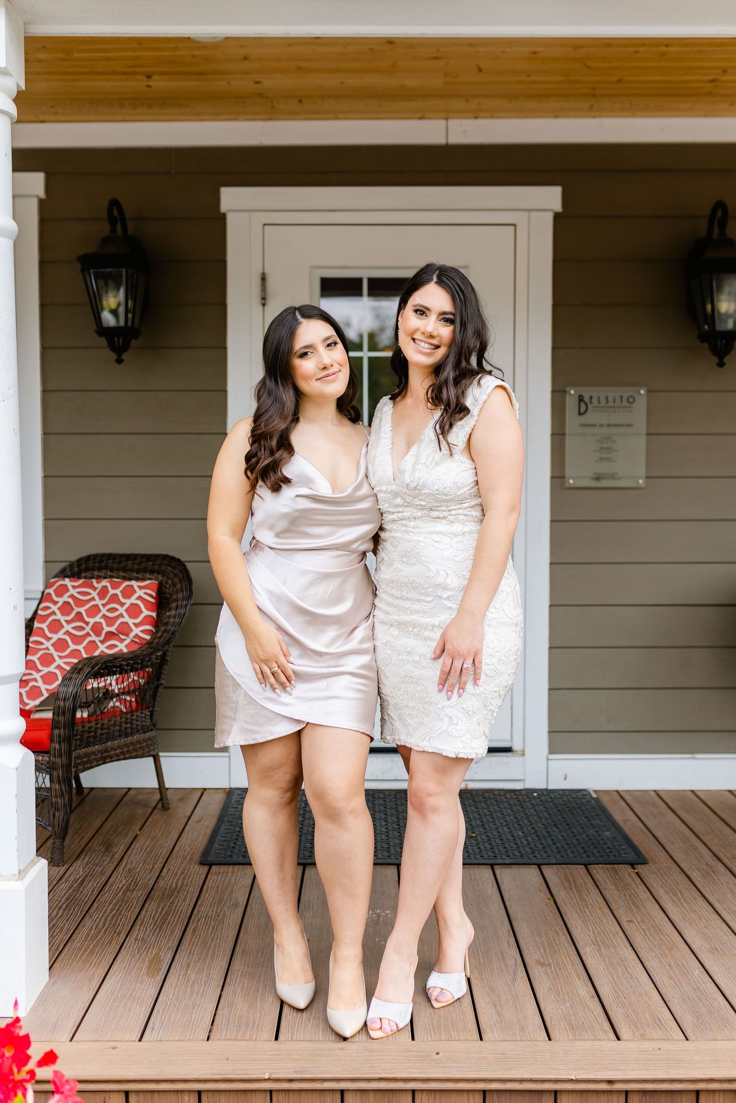 Two women in light colored dresses posing together on a porch with lanterns and red cushions.