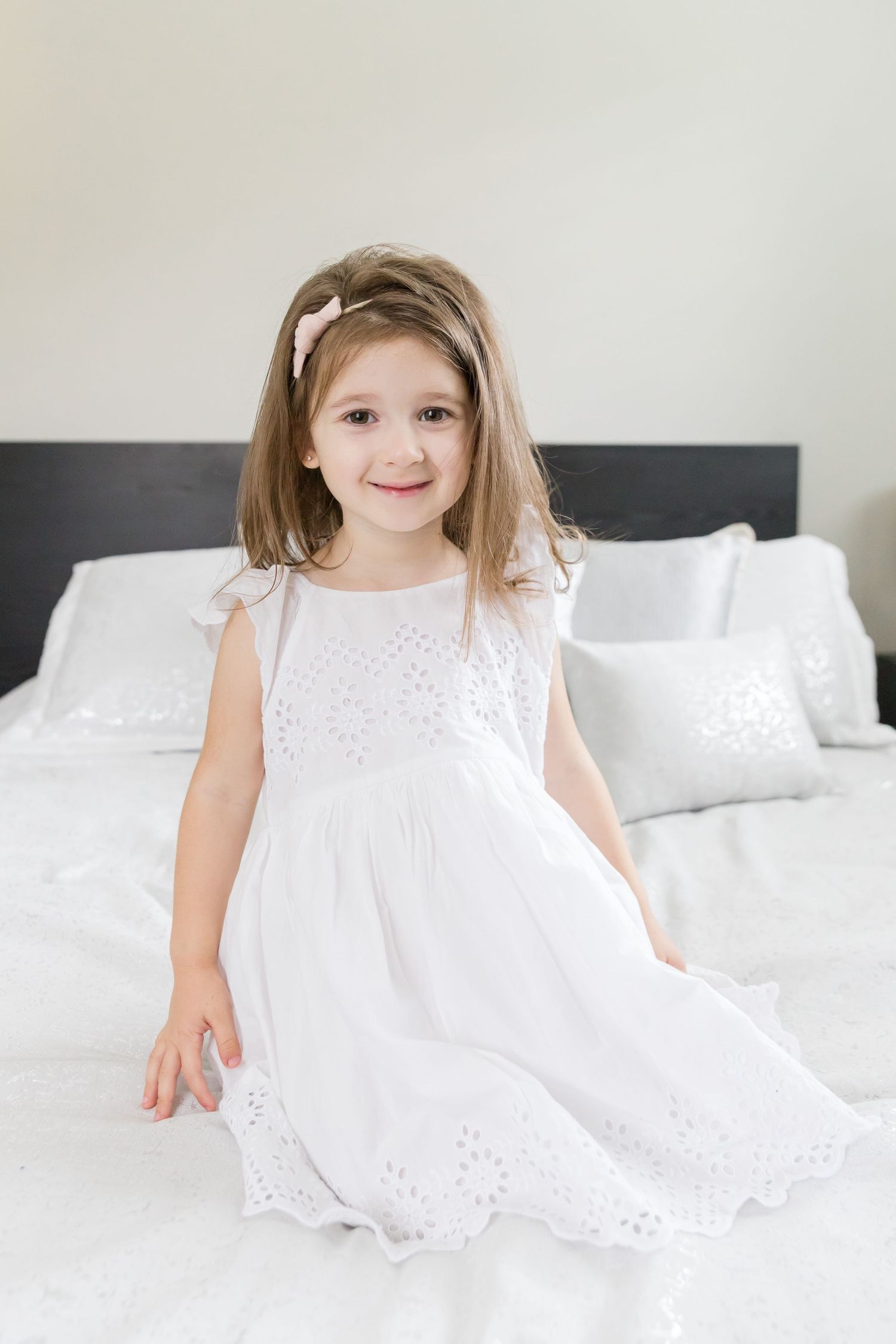 Child wearing white eyelet dress poses sweetly on white bed.