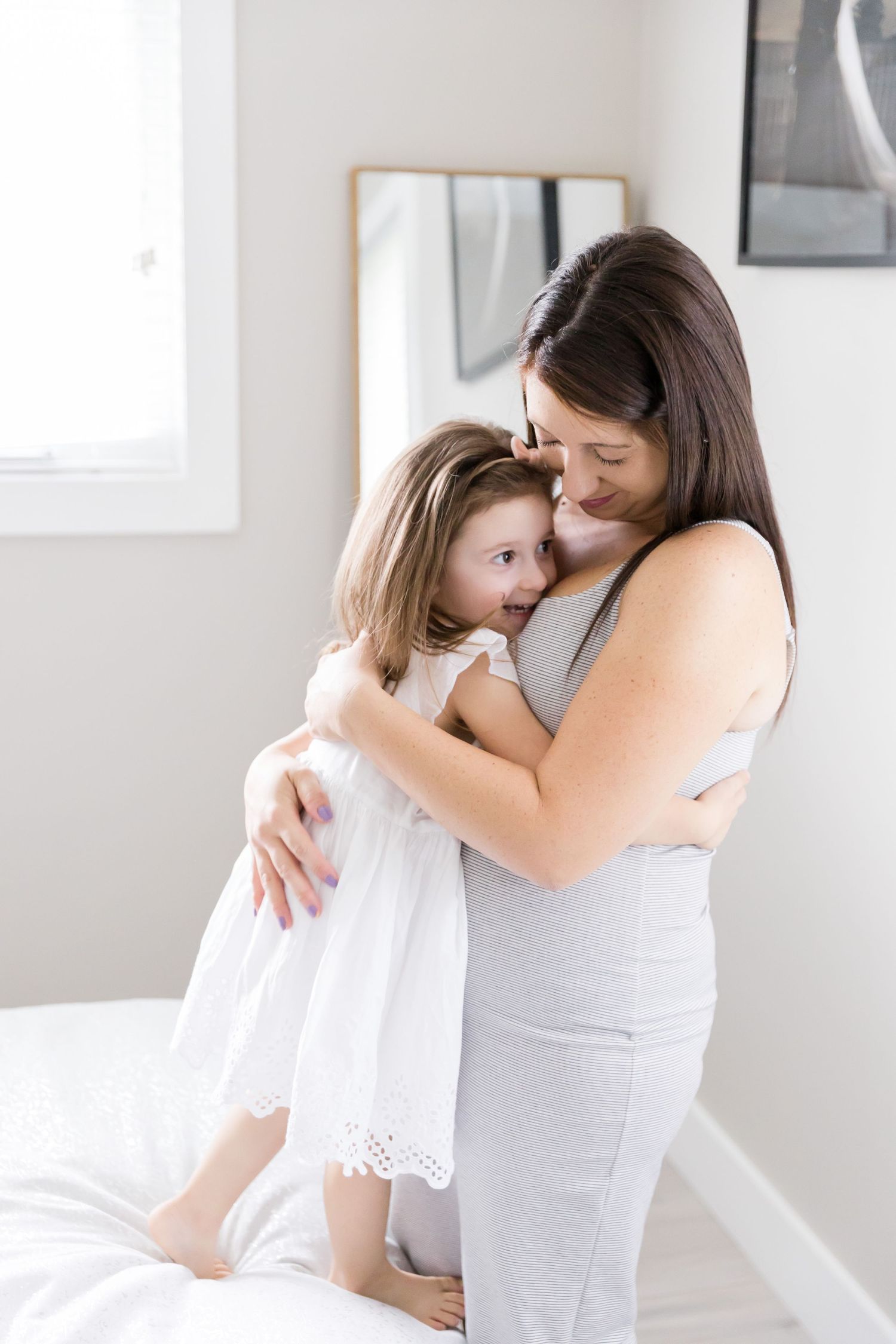 Tender moment between mother and child in white hugging against grey wall.