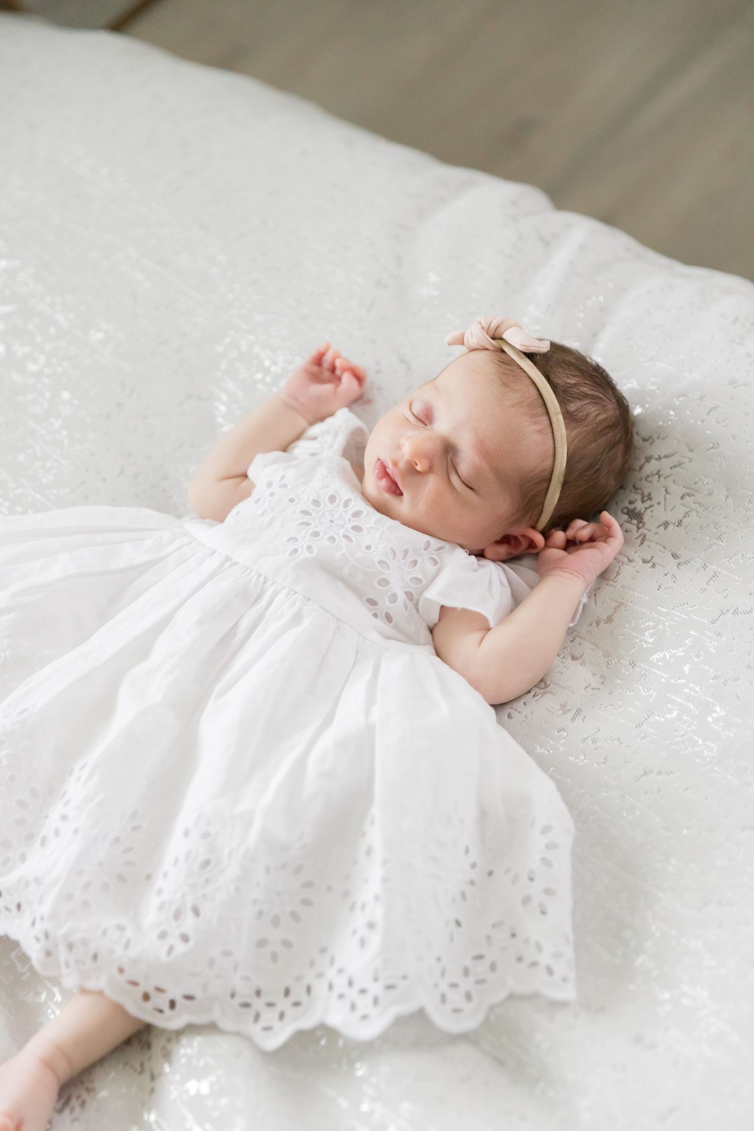 Sleeping baby in delicate white dress with headband lying on white bedding.
