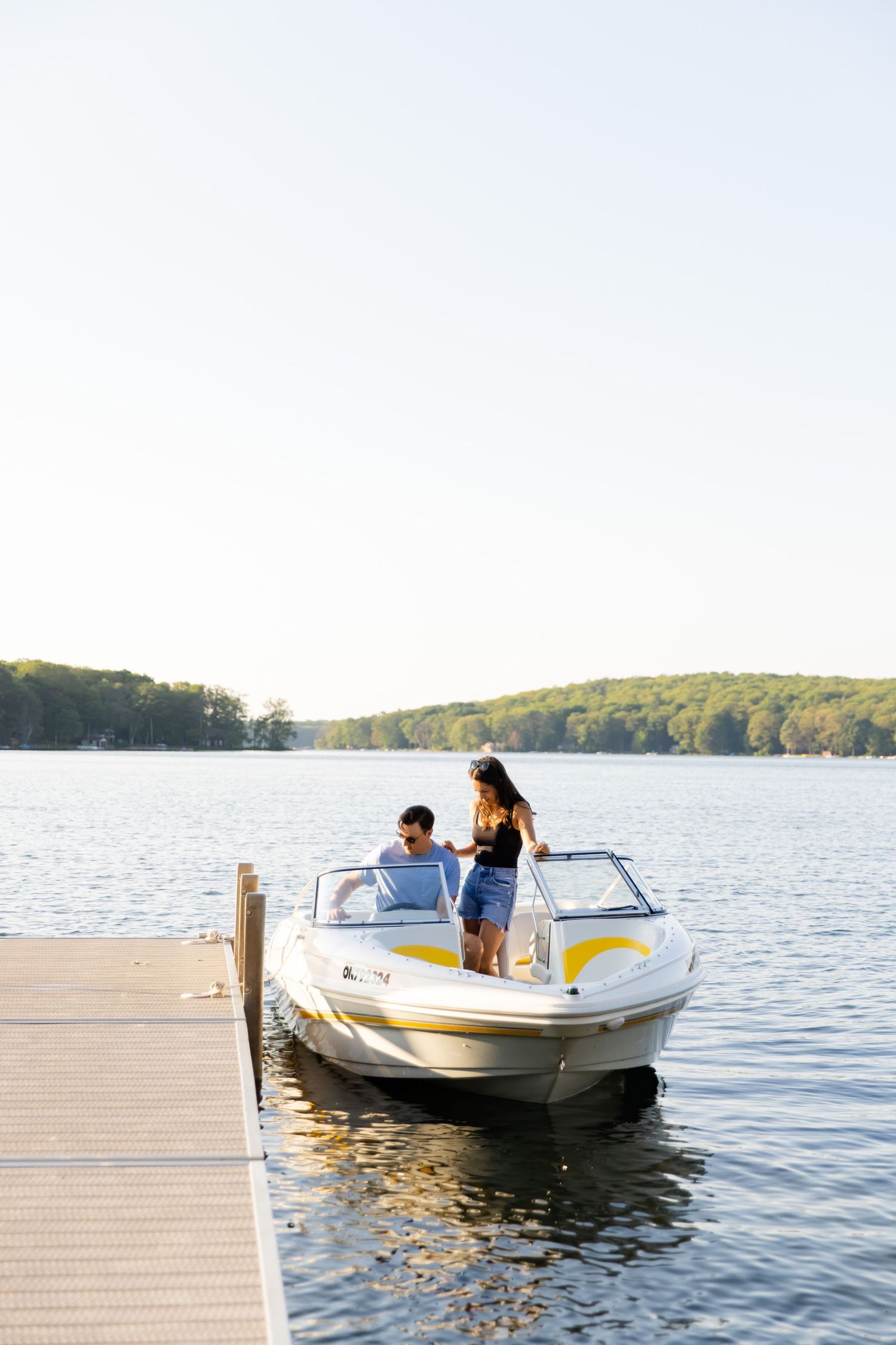 Couple enjoying a sunny day on a boat docked at a wooden pier on a tranquil lake surrounded by trees.
